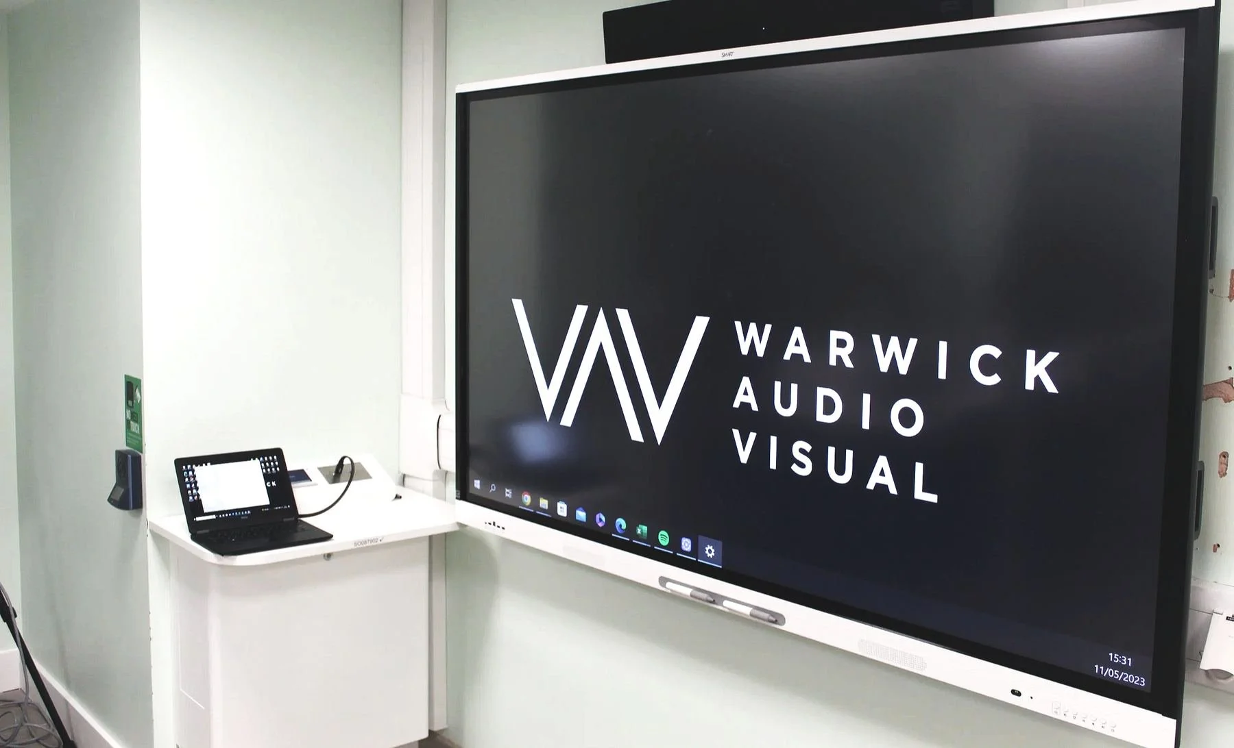 Classroom interactive display showing the Warwick Audio Visual logo, with a laptop on a lectern beside it