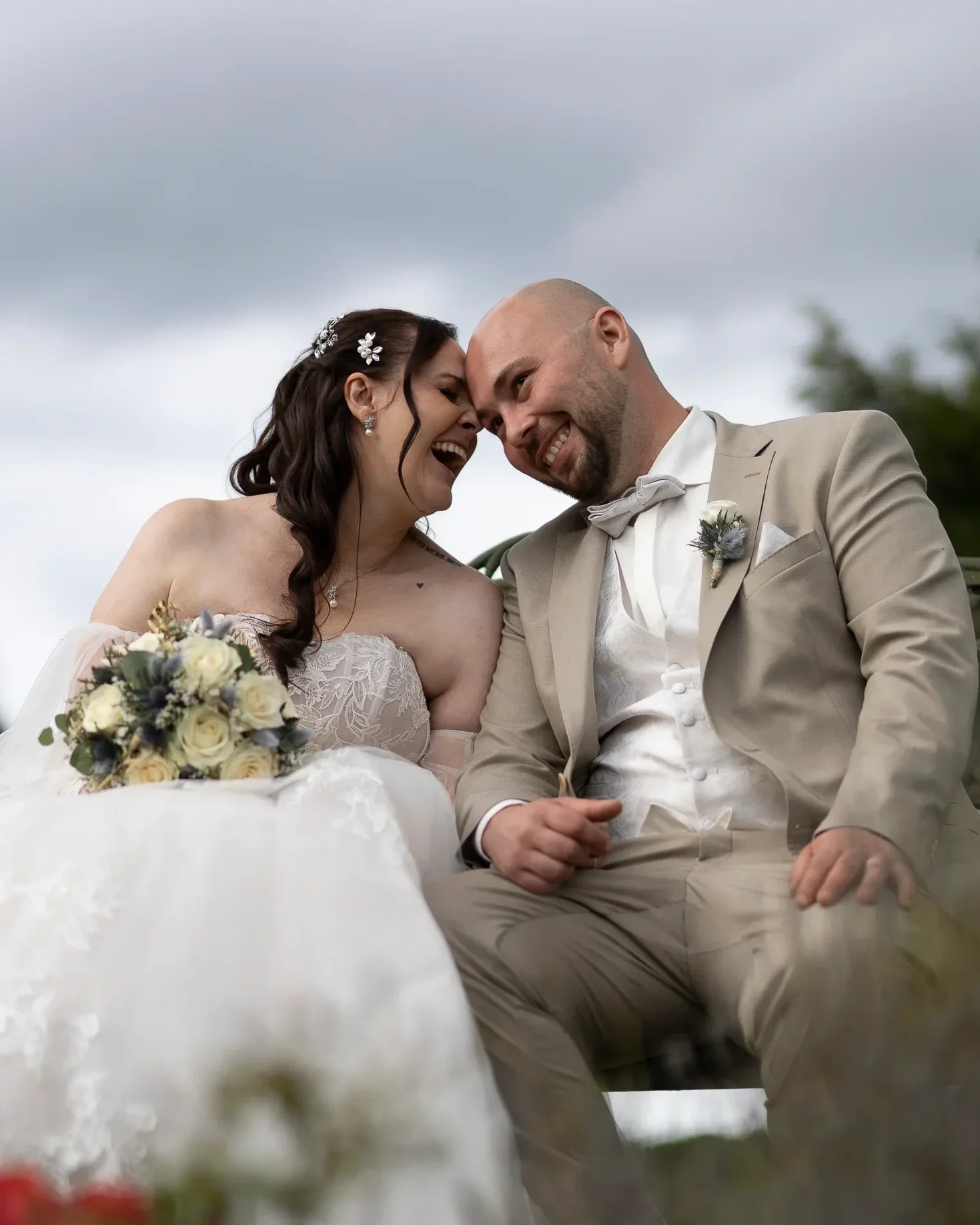 Ein glückliches Brautpaar bei der Hochzeit, sie lachen gemeinsam eng beieinander, er trägt einen hellbeigen Anzug mit Fliege, sie trägt ein weißes Brautkleid mit Spitze und Haarschmuck, im Hintergrund bewölkter Himmel.