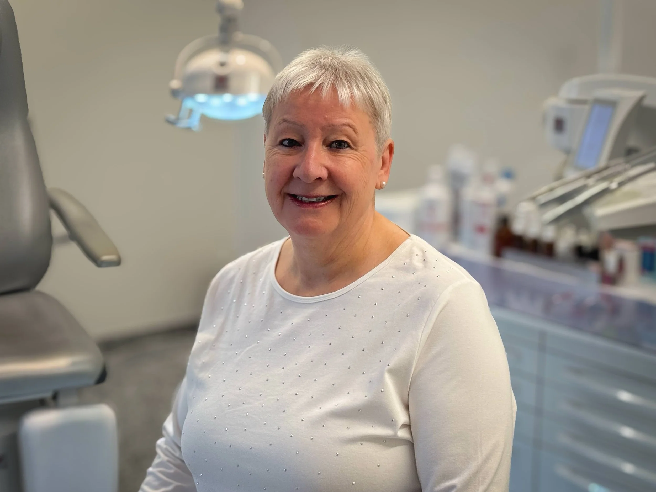 Eine Frau mit kurzen grauen Haaren in einem weissen Langarmshirt, sitzt in einer Podologiepraxis und lächelt in die Kamera.