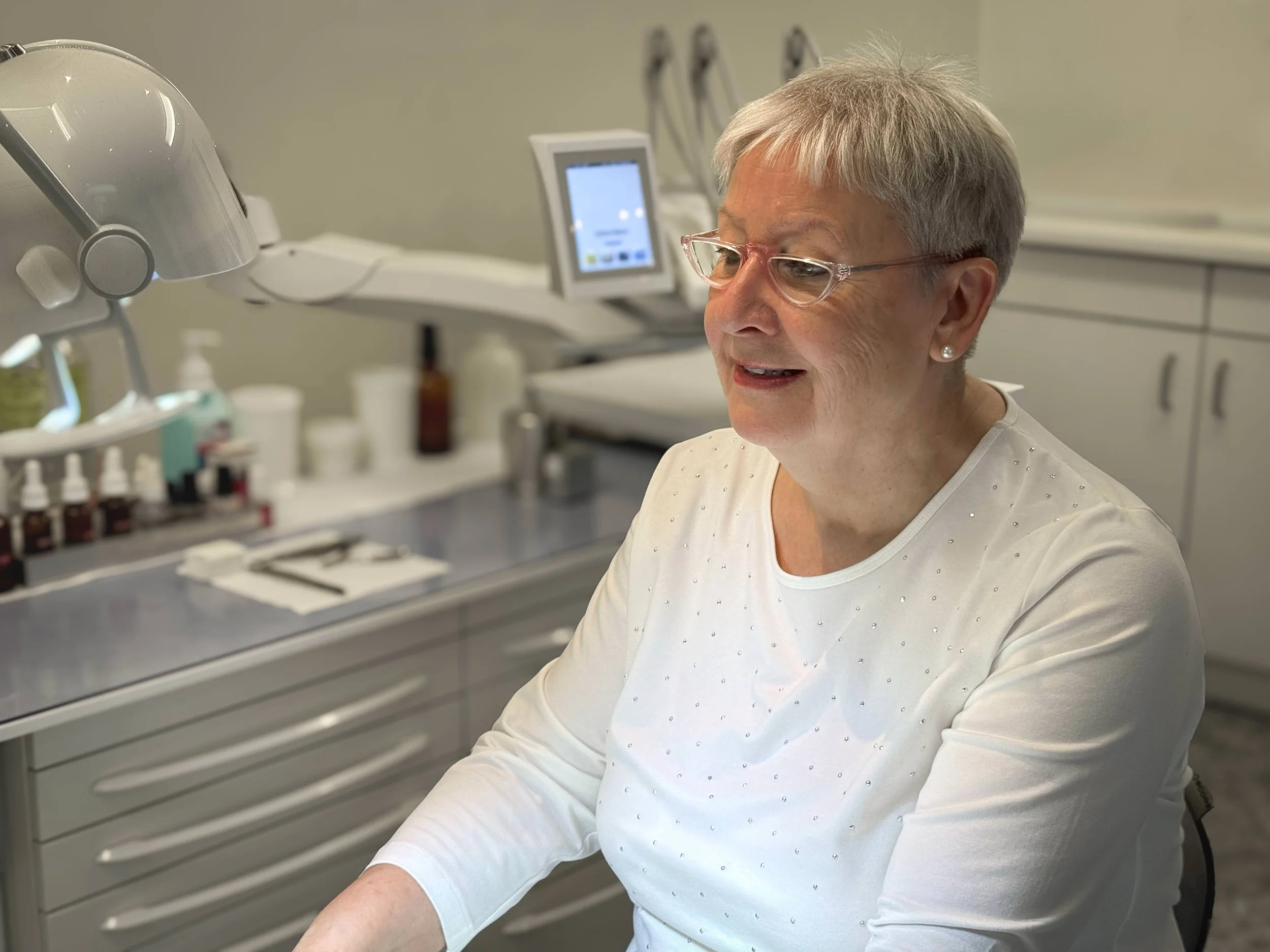 Frau in einem medizinischen Behandlungsraum, sitzend und lächelnd, mit medizinischer Ausrüstung im Hintergrund.