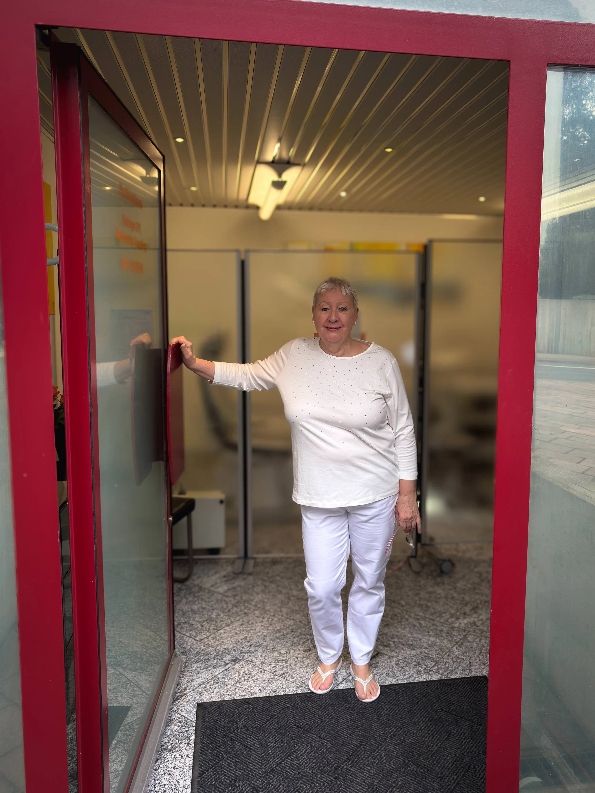 Frau in weißer Kleidung steht an der Tür einer Podologiepraxis, lächelt und blickt in die Kamera.