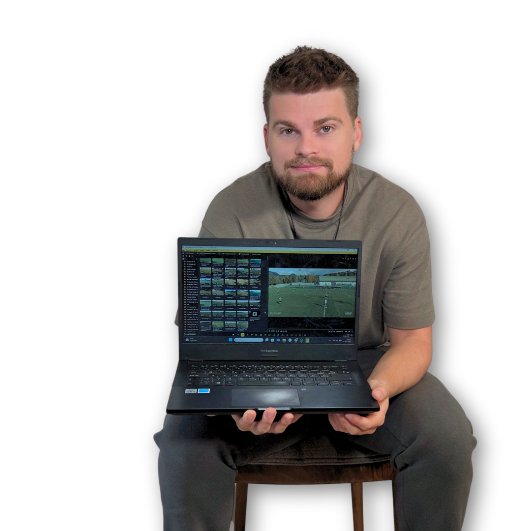 A young man with a beard and short brown hair, sitting on a wooden stool against a black background, holding an open laptop displaying a video editing or photo editing software.