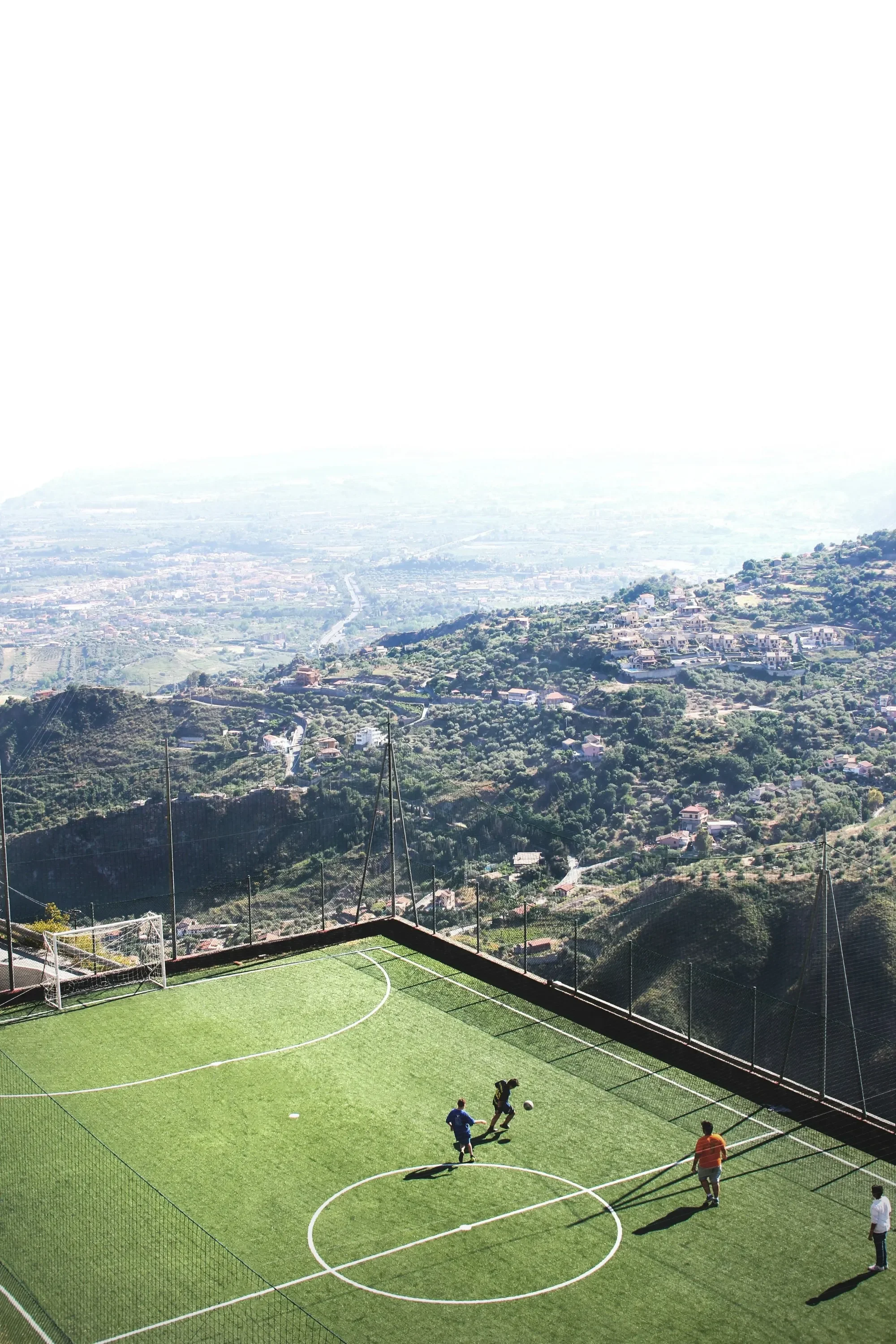 People playing soccer on a small artificial turf field on a hillside with a scenic view of the valley and mountains in the background.