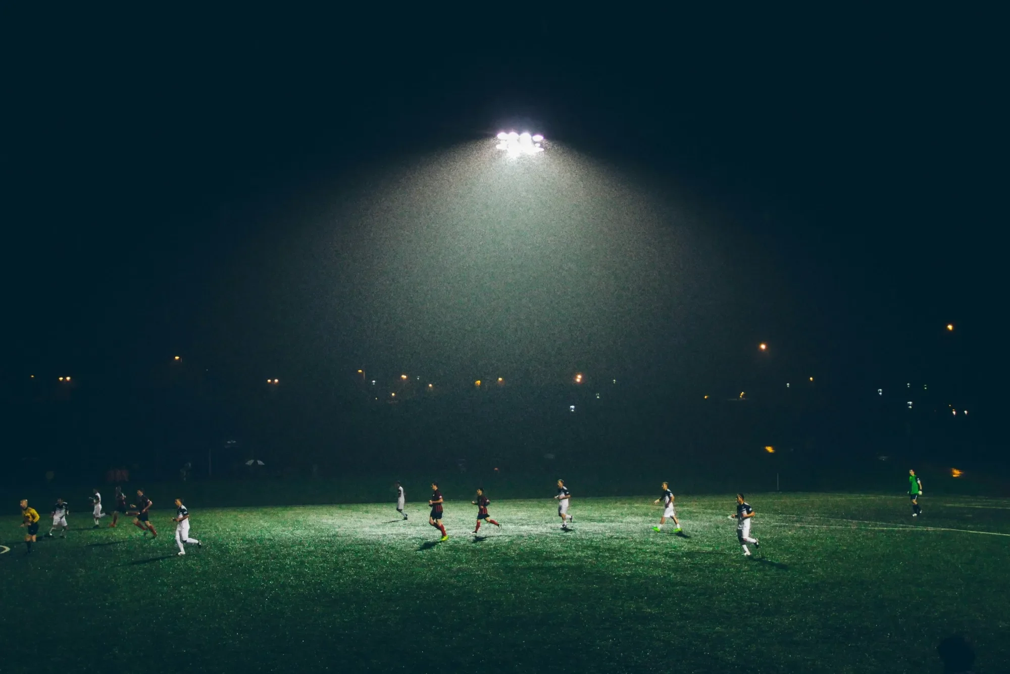 Soccer players playing under stadium lights during a night match on a rainy evening.