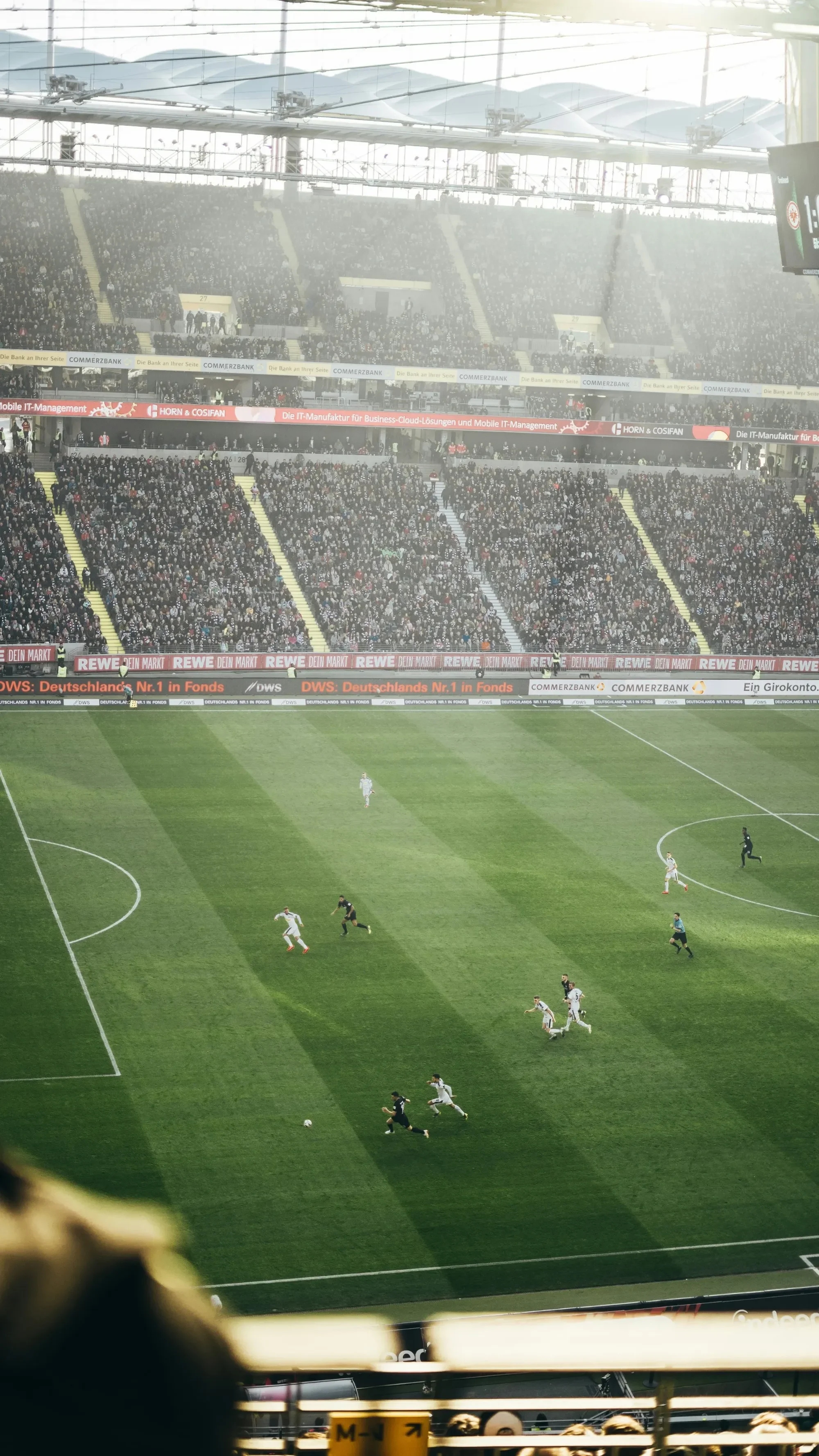 A soccer match taking place in a large stadium. Players are running on the field, with some chasing the ball. The stands are filled with spectators, and advertising banners line the perimeter of the field.