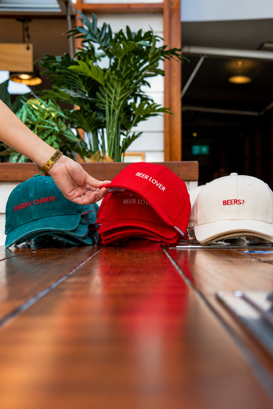 Several branded caps with humorous embroidered text on a wooden table, with a person's hand adjusting one of the red caps. The caps have phrases like "CHEEKY CHERRY," "BEER LOVER," and "BEERS?" against a background of plants and an indoor setting.