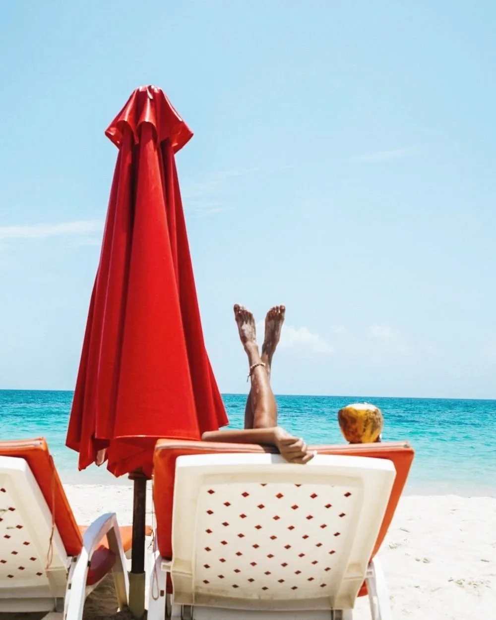 Person relaxing on a beach lounge chair with legs raised, next to a coconut on the sand, under a closed red beach umbrella, facing the ocean with clear blue sky.