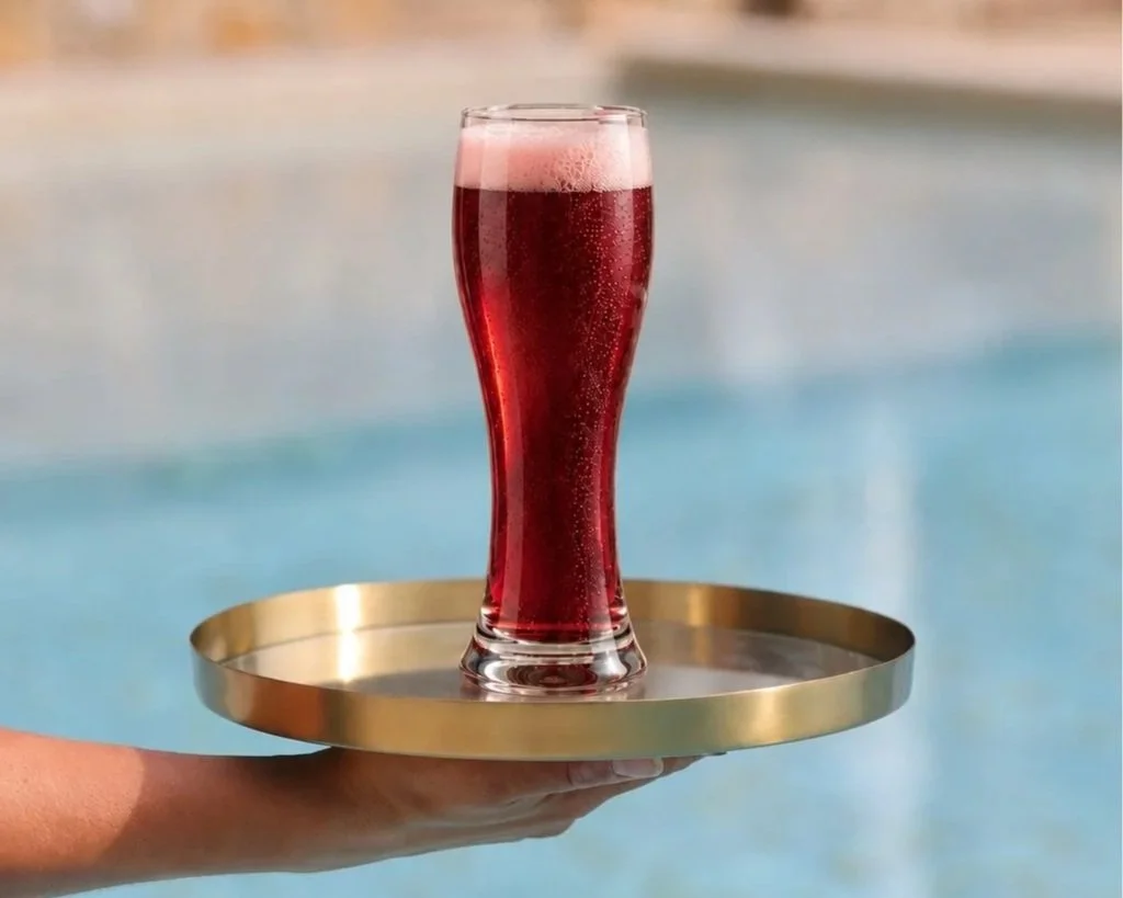 A tall glass of Gold Coast Cherry beer and pinkish head on top is being served on a round metal tray by a person at a poolside area.