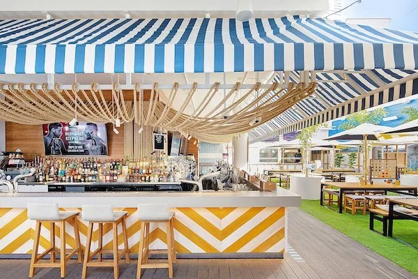 Outdoor bar area with striped blue and white awning, wooden ceiling decor, and seating including barstools and tables with umbrellas. Surfers Paradise Beergarden Gold Coast venue 100 years