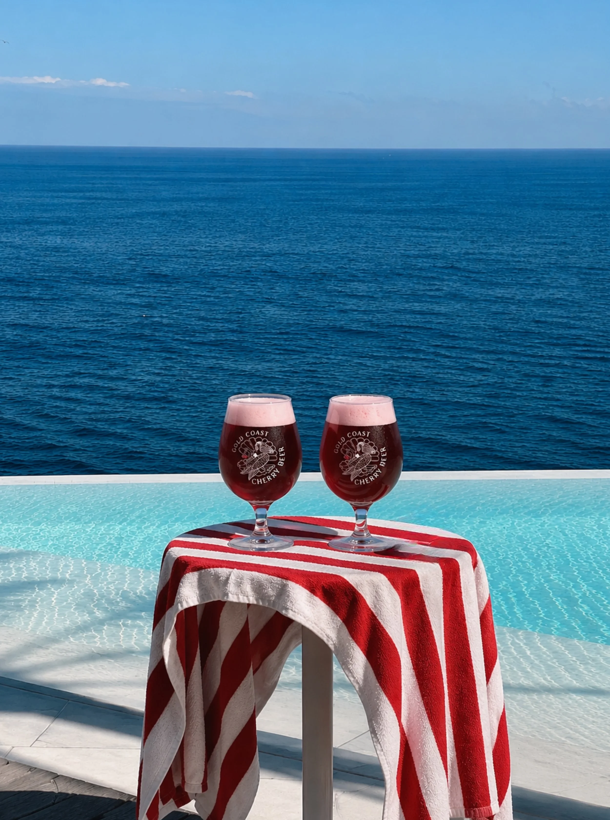 Two glasses of cherry beer on a striped towel, with the ocean in the background and a clear sky.