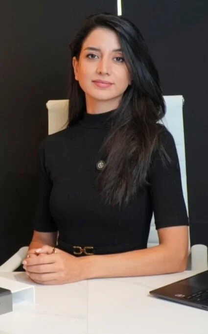 A woman with long dark hair wearing a black top, sitting at a white desk with a laptop in front of her, in an office setting.