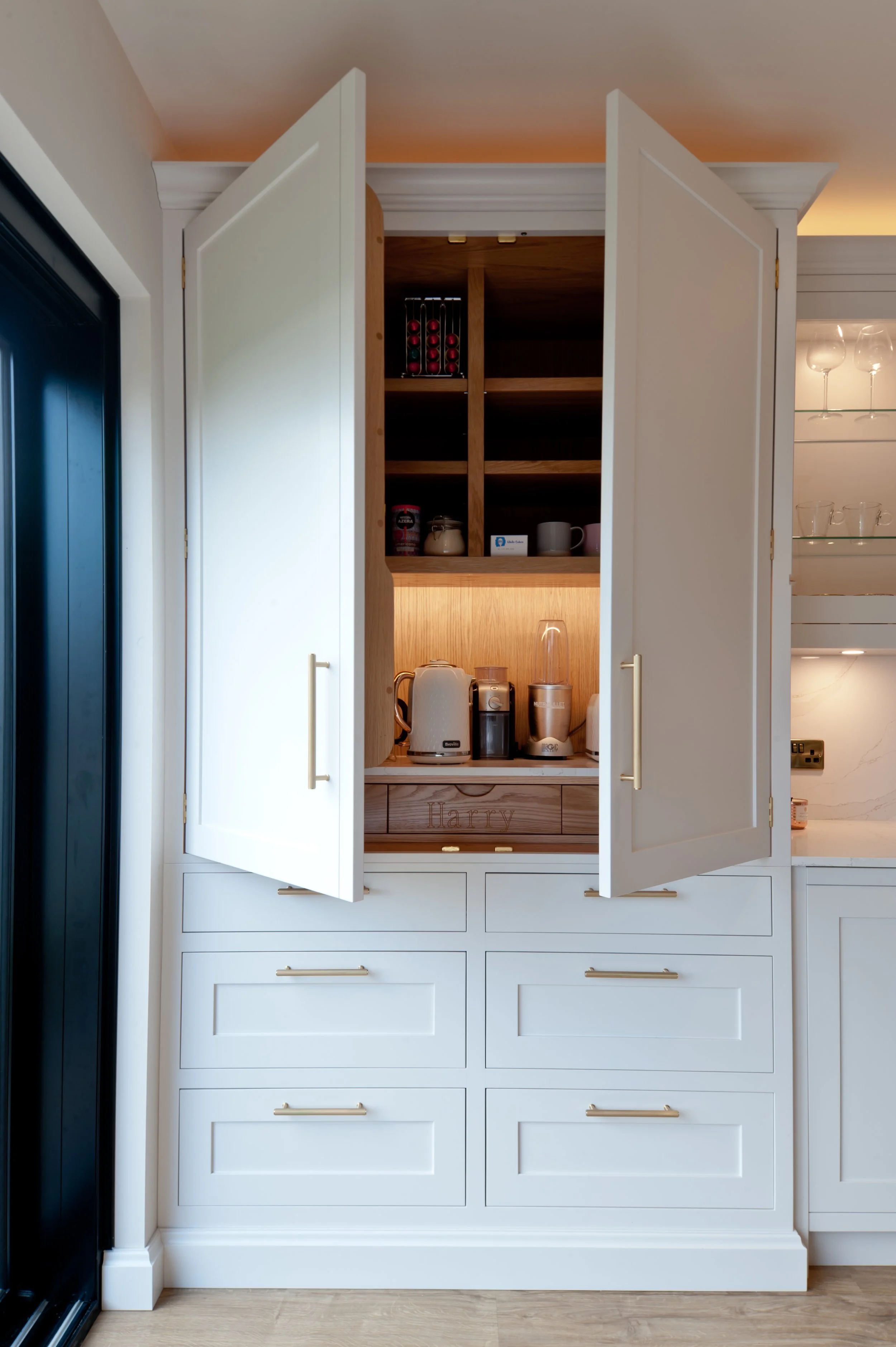 White kitchen cabinet with open doors revealing shelves and kitchen items inside, set against a white wall with wooden floors.