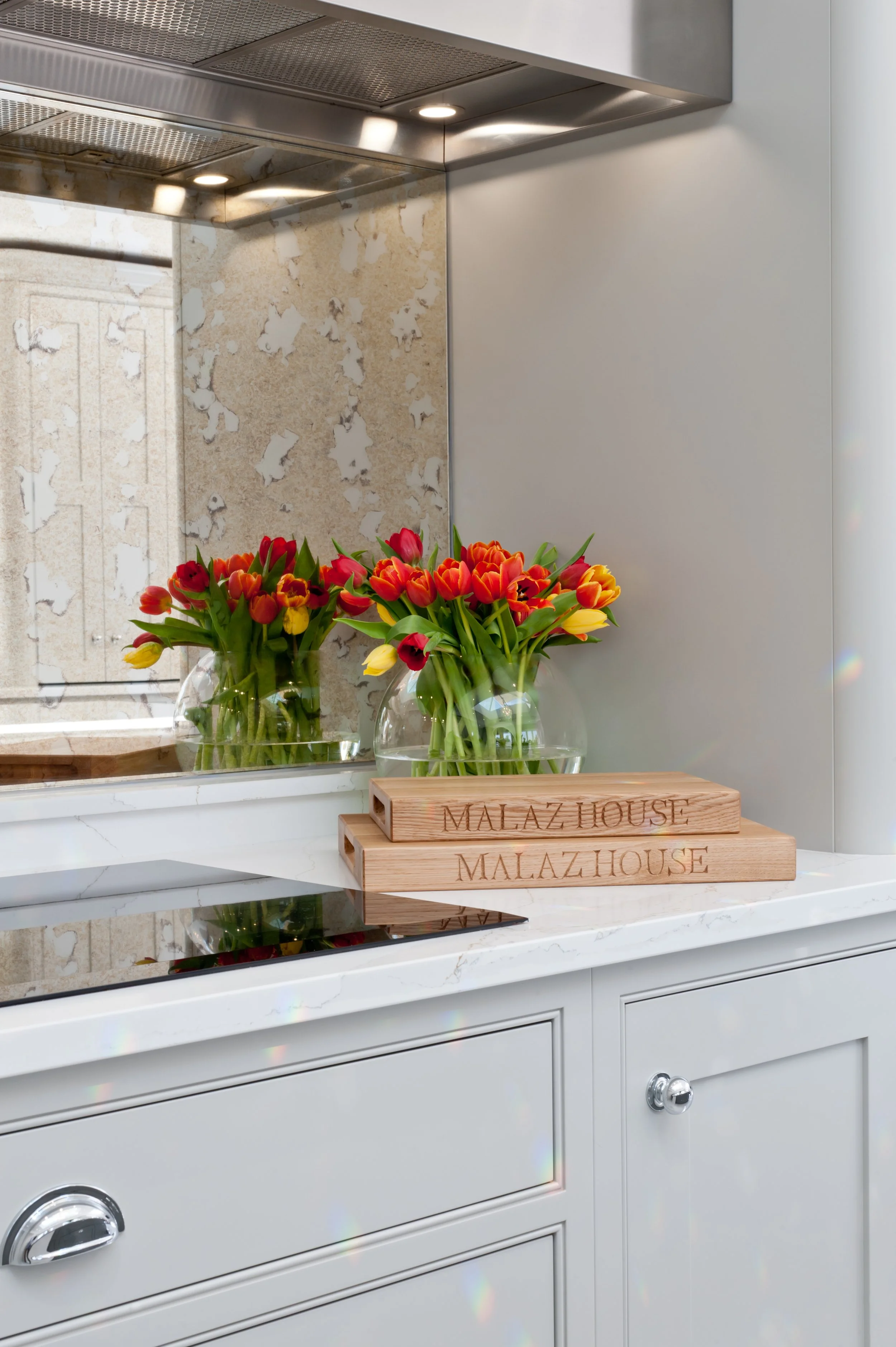 A white kitchen countertop with a glass vase of red, yellow, and orange tulips. Next to the vase are two wooden boxes with the words 'MALAZ HOUSE' engraved on them. A reflective backsplash and modern stainless steel range hood are visible in the background.