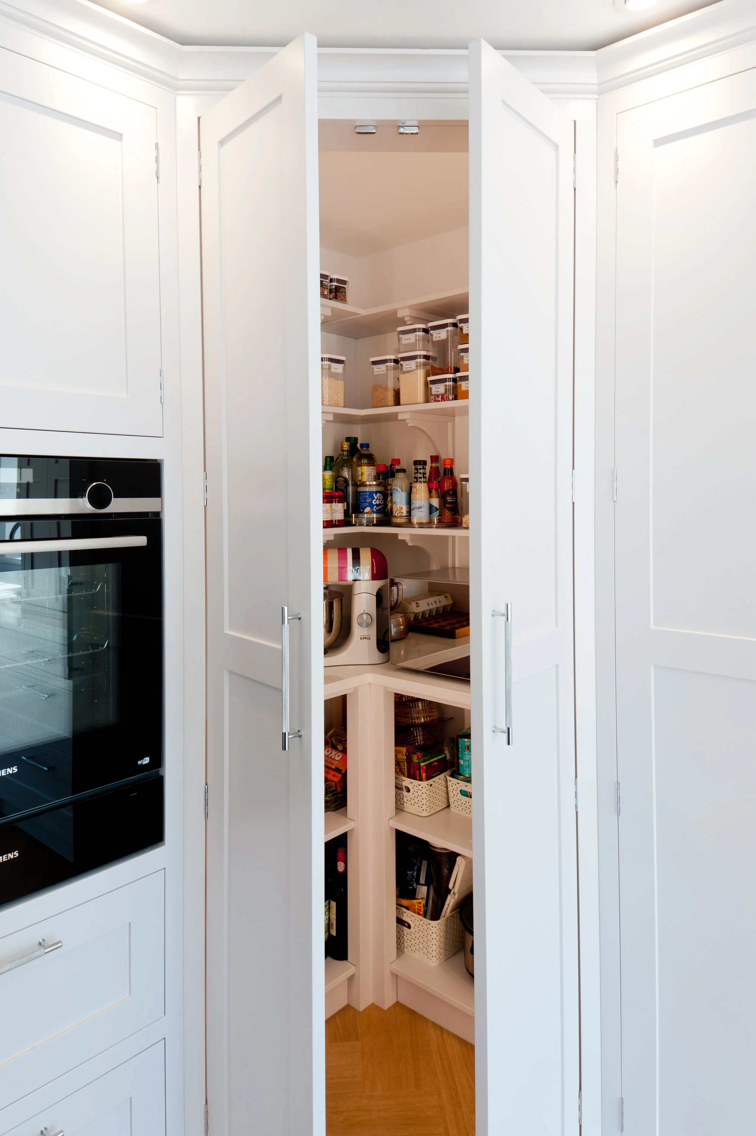 Kitchen pantry with white shelves and various food items and condiments inside, located between white cabinetry. To the left, there is a built-in oven with a glass door.