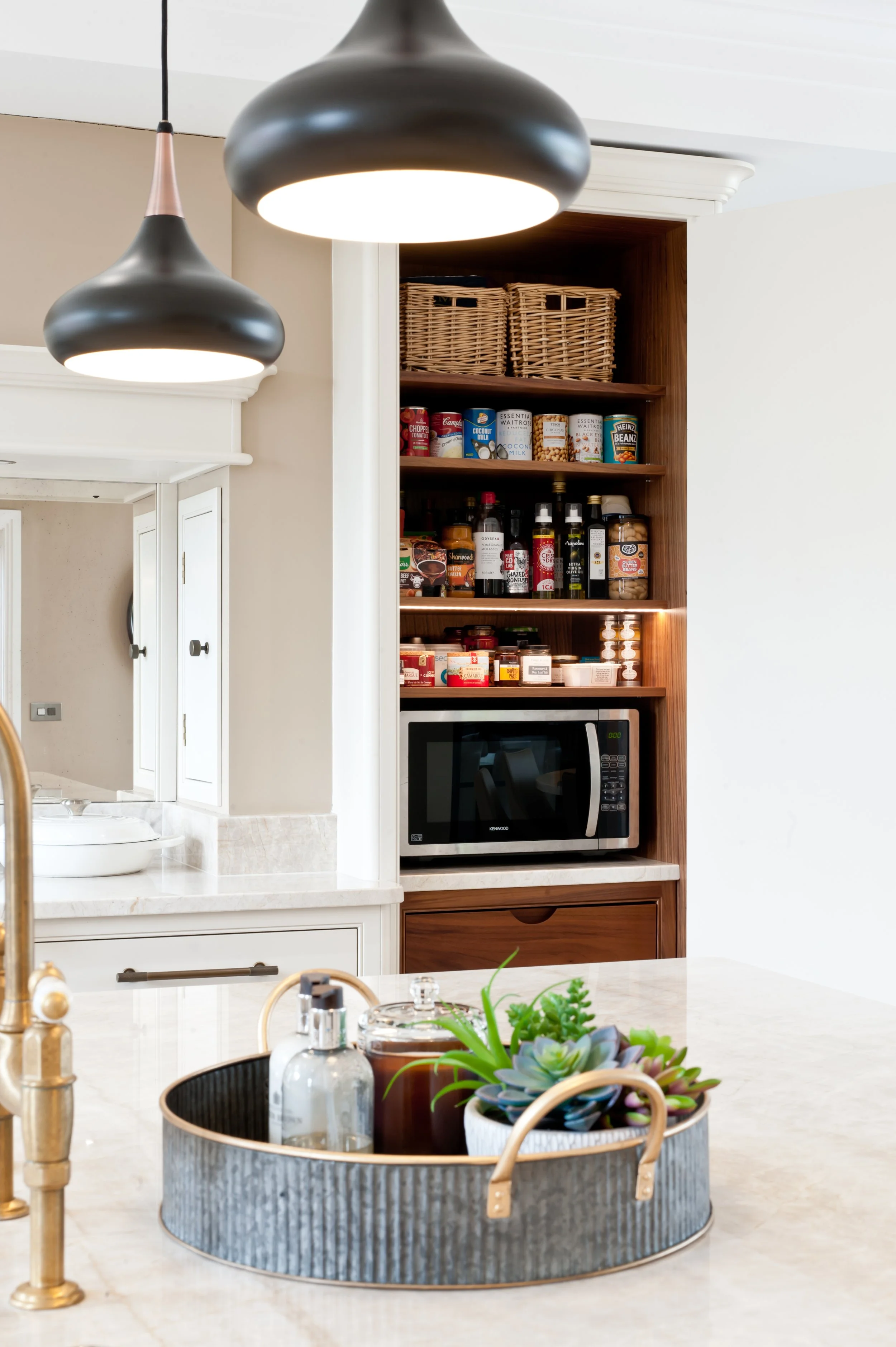 View of kitchen cabinetry with open pantry shelf containing woven baskets, canned goods, bottles, and jars, adjacent to microwave oven. In the foreground, a kitchen island with a plant in a basket, glass bottles, and dental floss container, and overh