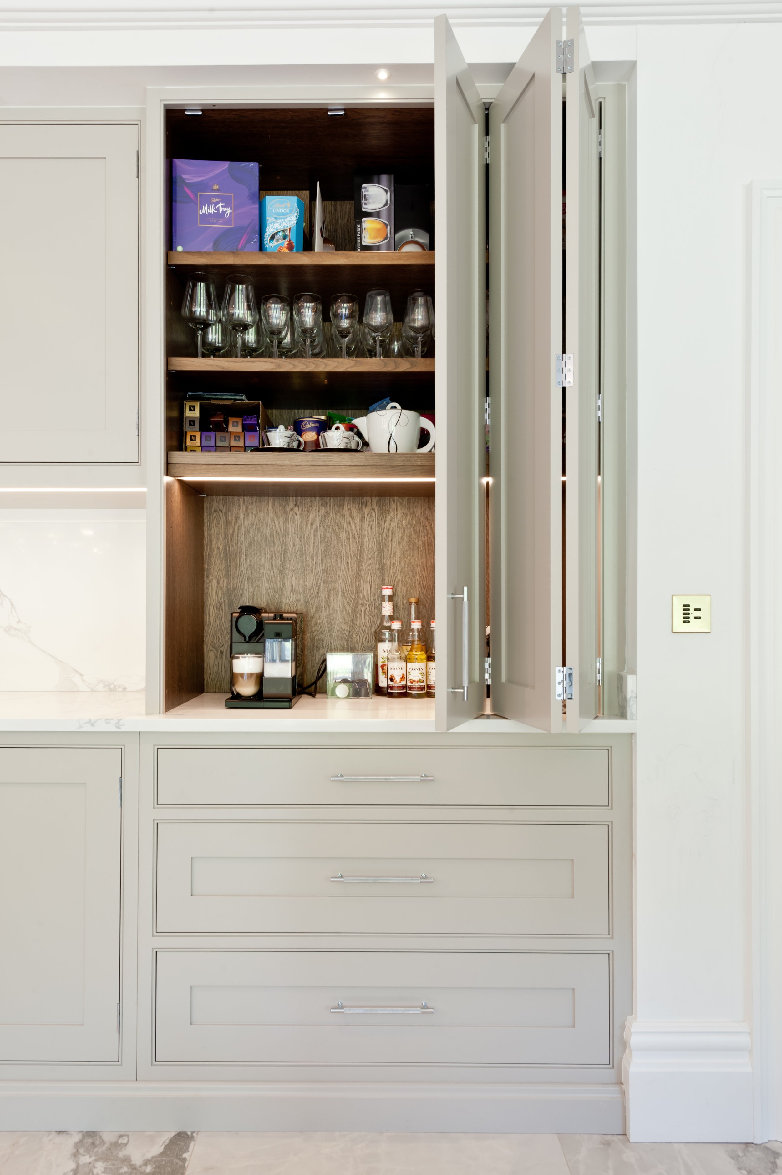 Built-in way with open doors revealing shelves with glassware, drinks, mugs, and snacks inside a kitchen cabinet.