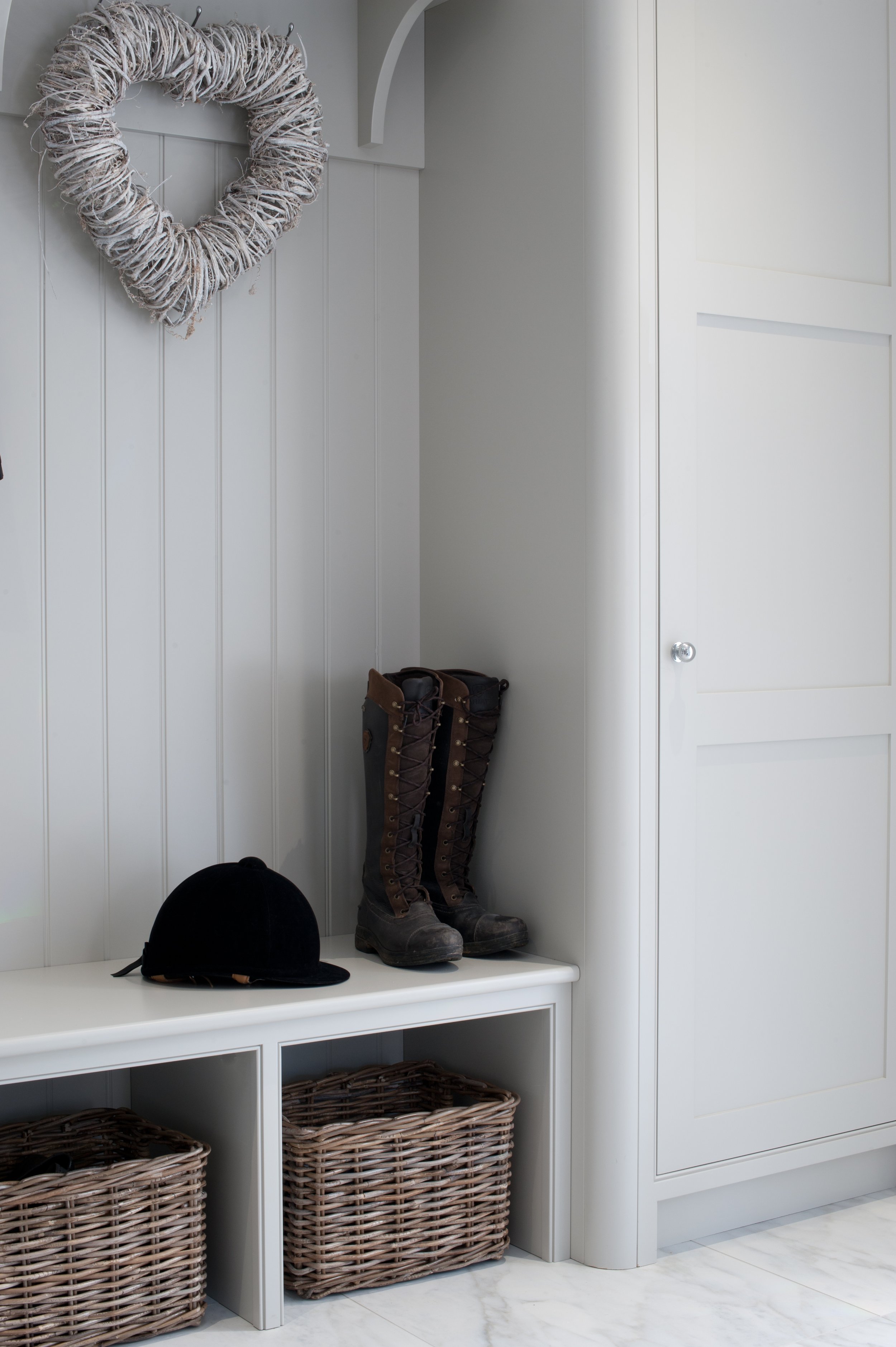 Bespoke white boot room cabinetry with built-in bench seating, wicker storage baskets, tall cupboards and space for riding boots and equestrian accessories.