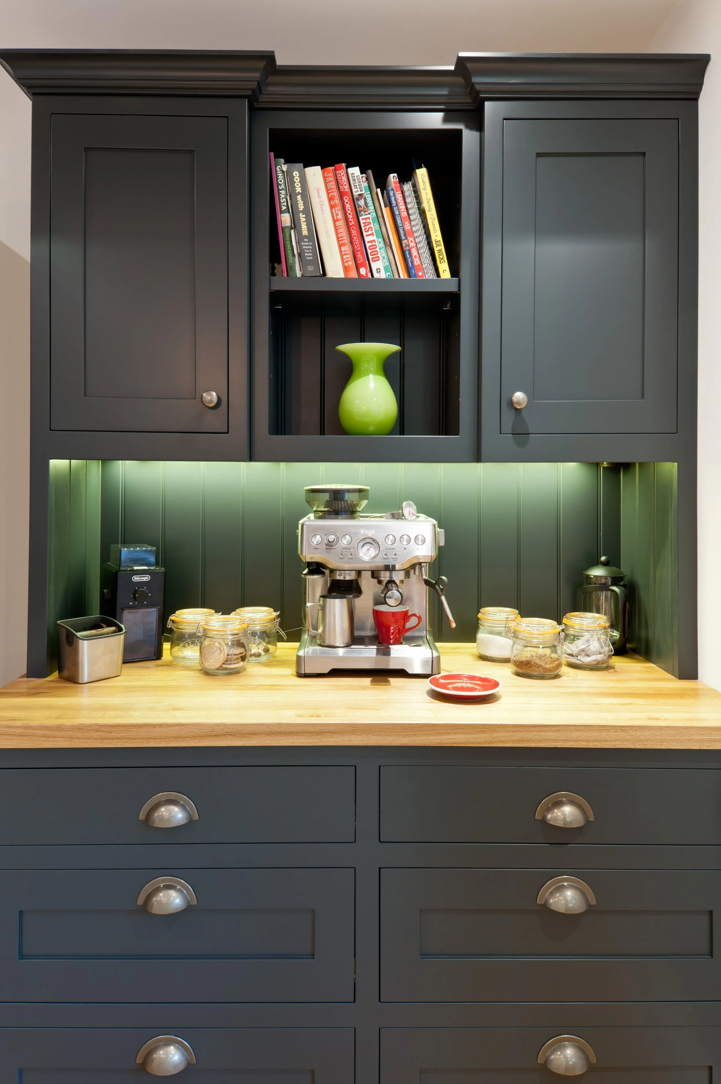 A kitchen counter with a coffee machine, jars, a red cup, and various kitchen items, with dark gray cabinets above containing a selection of cookbooks and a green vase.