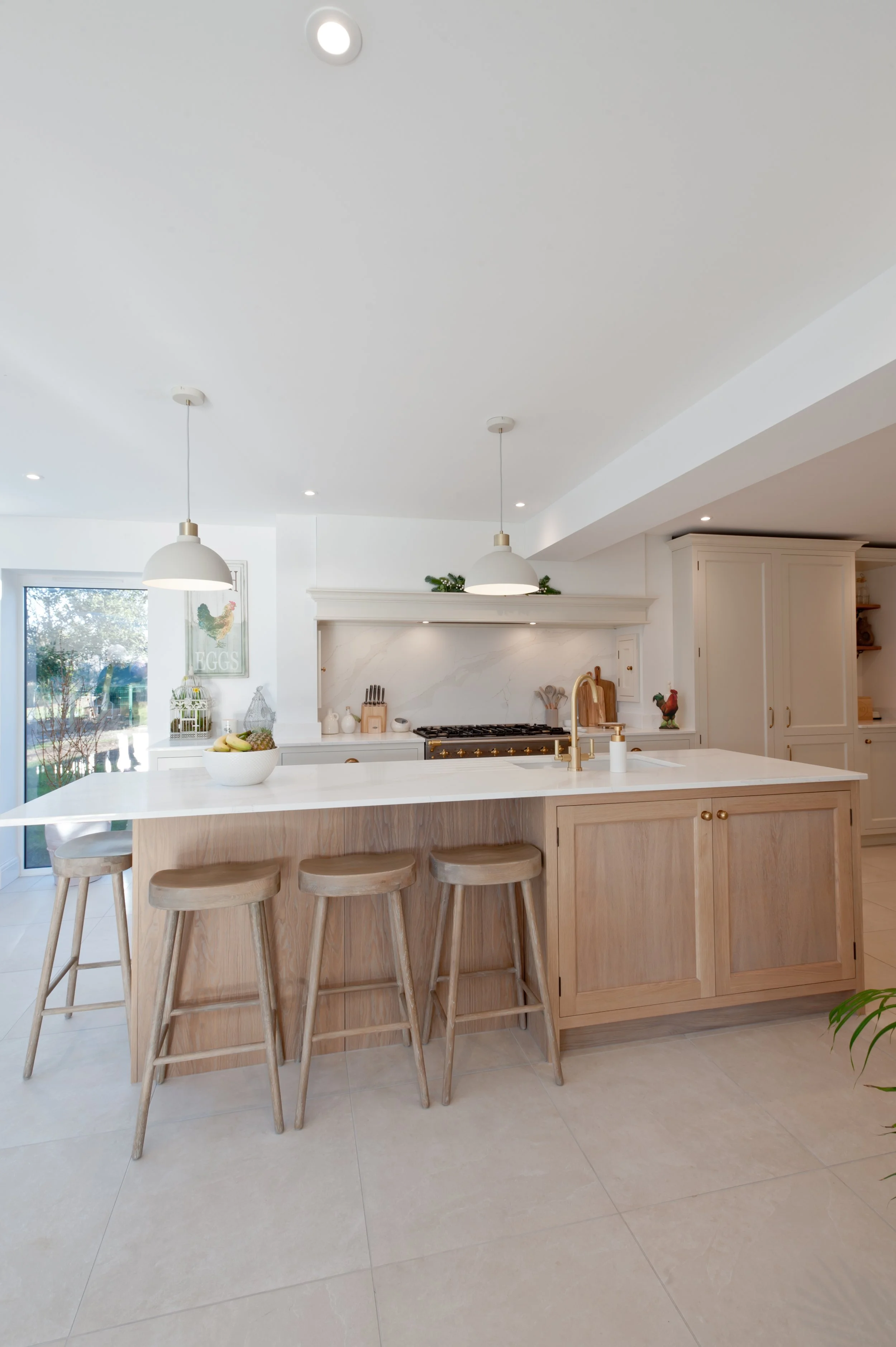 Modern kitchen with white walls and ceiling, wooden island with four stools, pendant lighting, and a window with a decorative chicken and 'EGGS' sign.