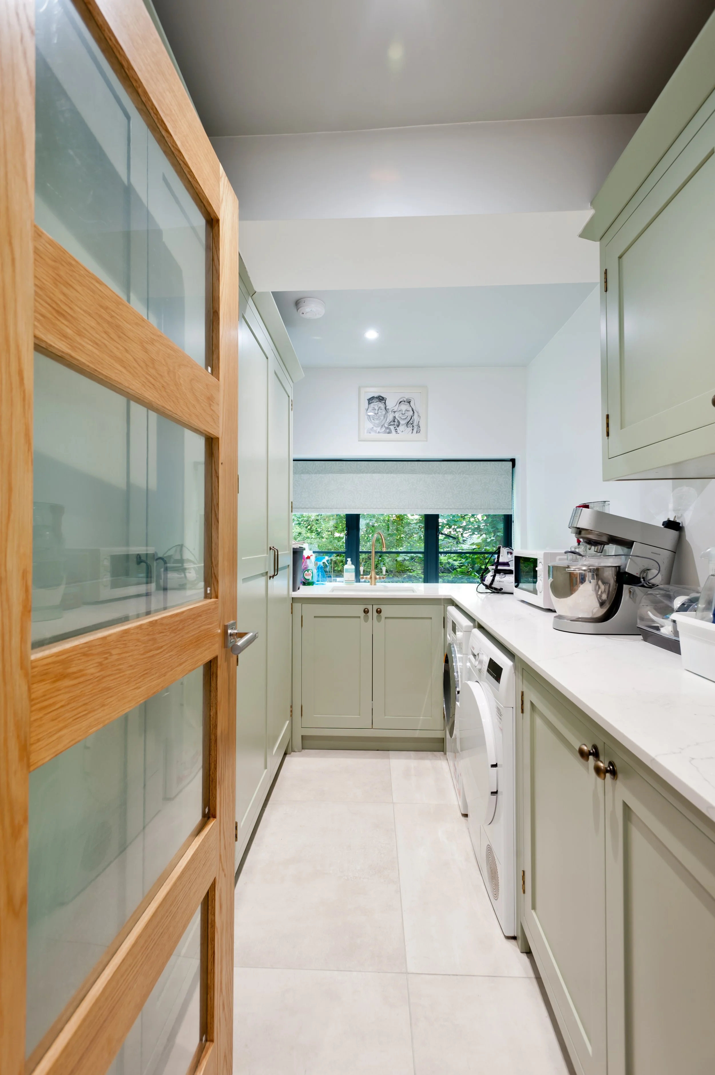 A view of a modern kitchen with green cabinetry, white countertops, a window with greenery outside, and various small appliances on the counter.