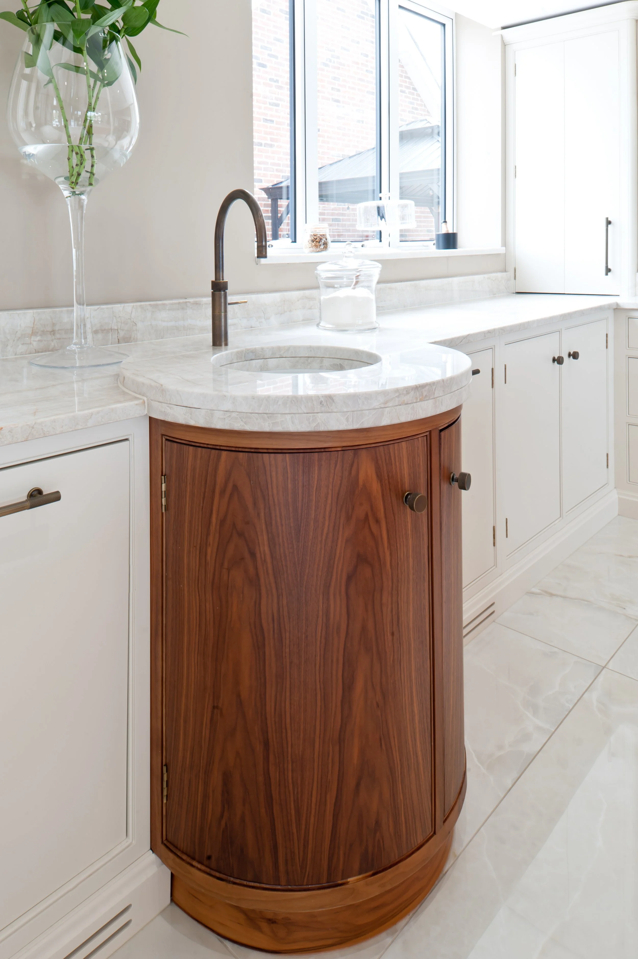 A kitchen with a marble countertop, a built-in sink with a bronze faucet, white lower cabinets with round handles, a small curved wooden cabinet, a large window, and various small decorative items on the windowsill.