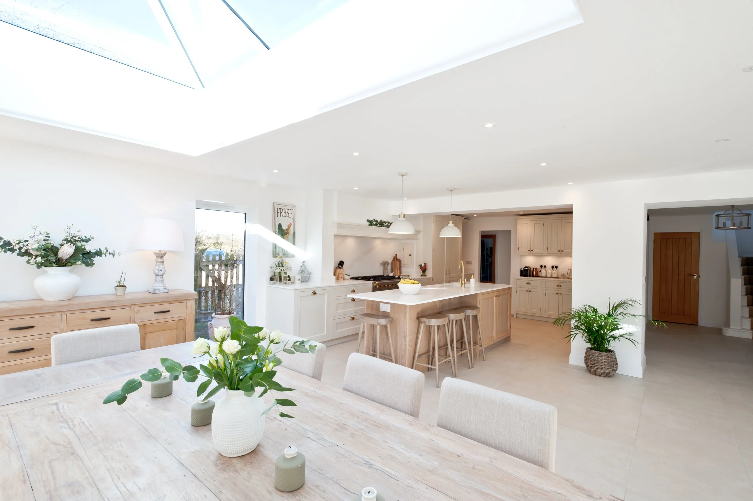 Bright, modern kitchen and dining area with white walls, light wood furniture, potted plants, and large skylight.