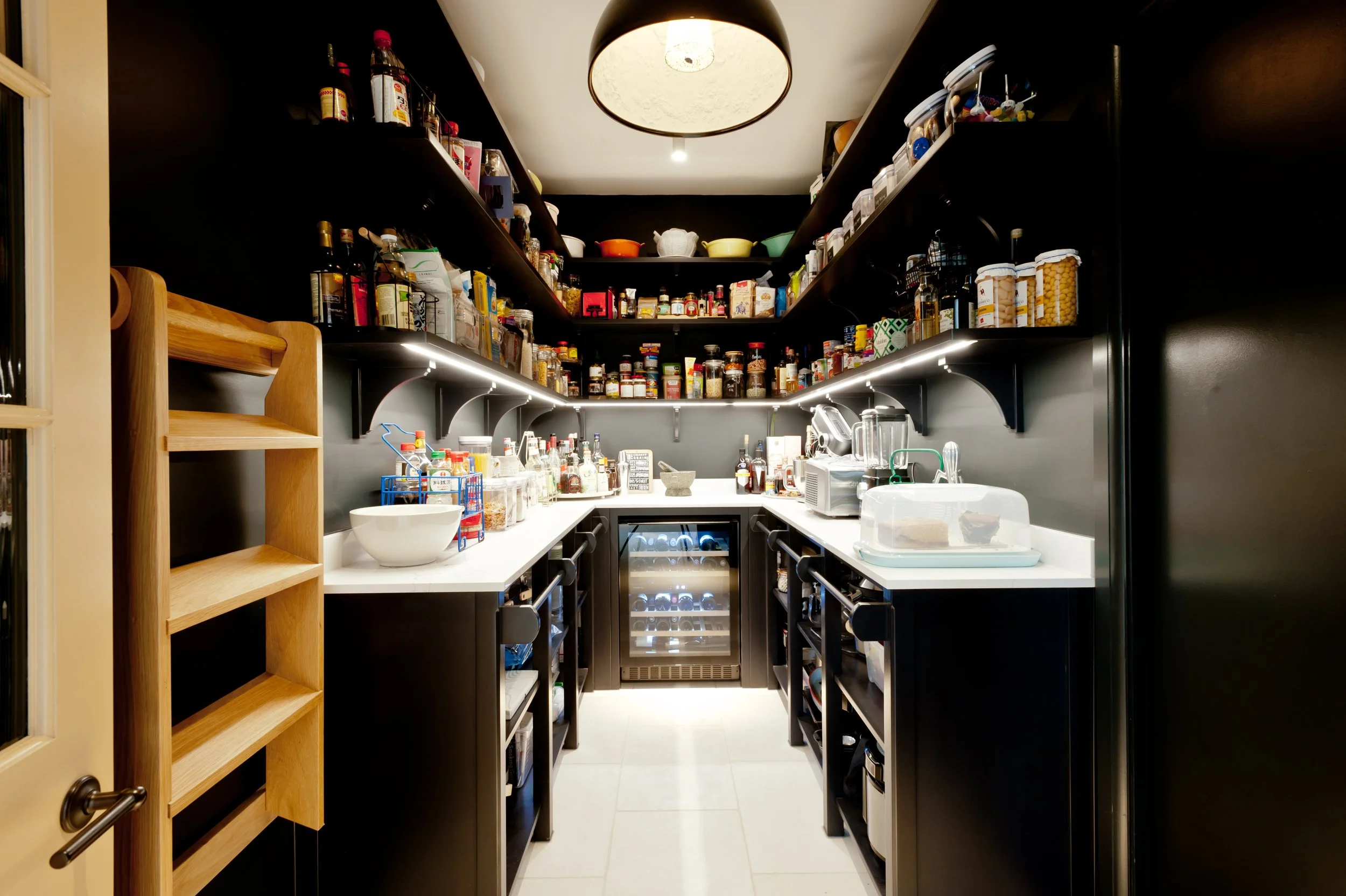A walk-in pantry with black shelves stocked with spices, jars, and kitchen supplies, and a white countertop with various kitchen items in front of black cabinets and a small wine cooler.