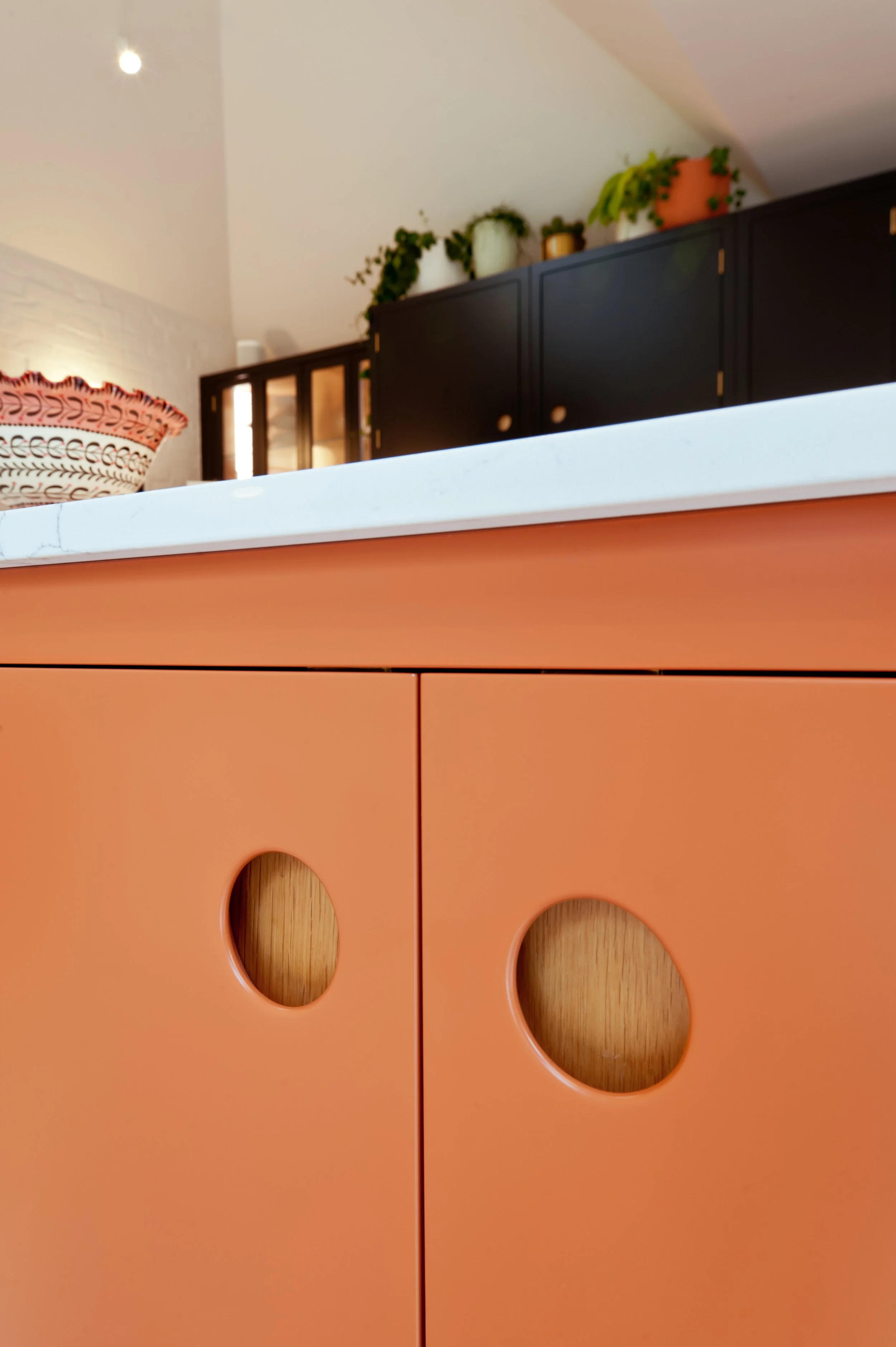 Close-up of a coral-colored cabinet with circular cutouts, with a white marble top. In the background, there are black cabinets and a white wall with shelves holding potted plants, and a decorative basket on the left side.