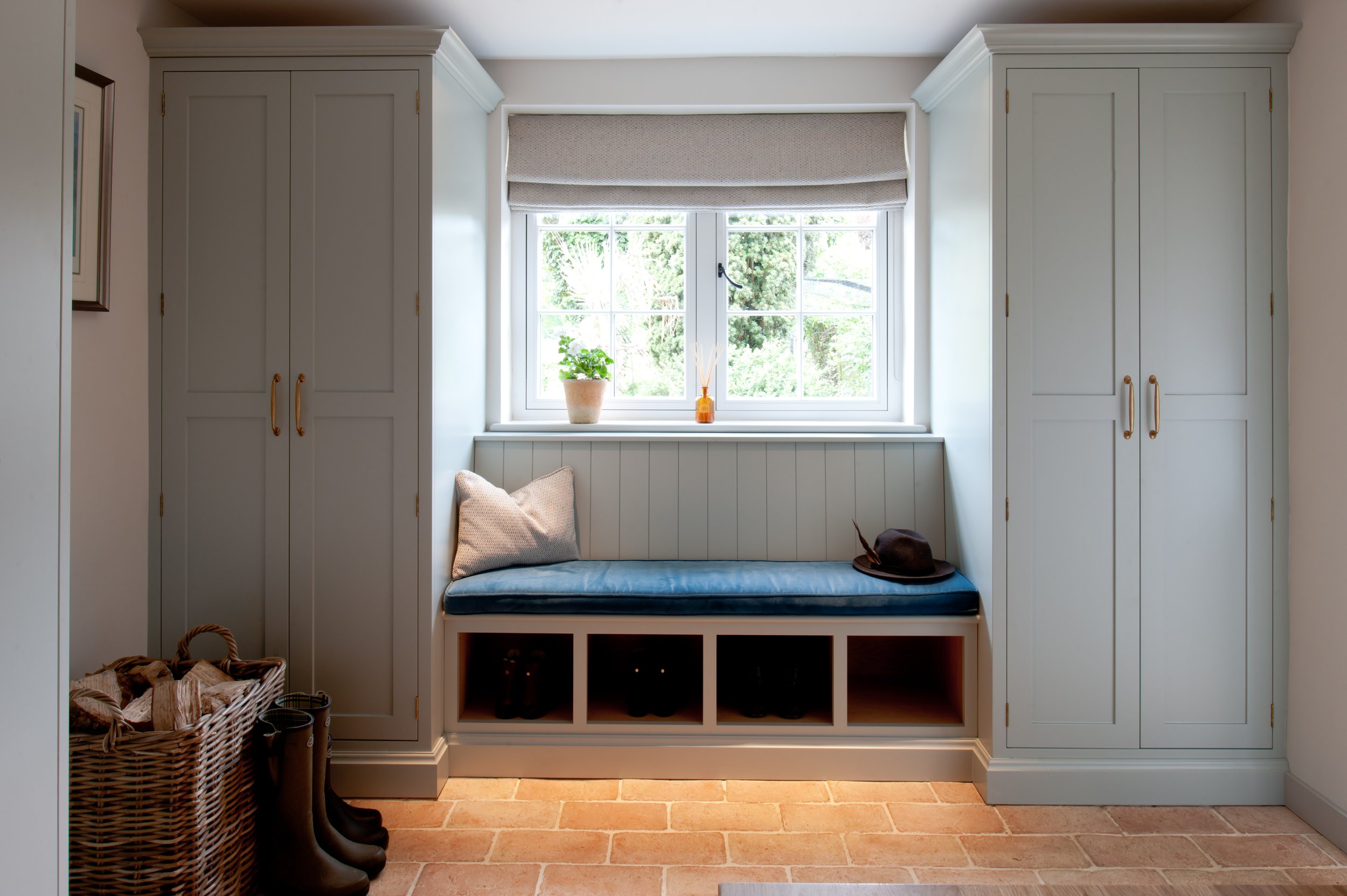 Beautifully proportioned bespoke boot room with symmetrical fitted cupboards, blue upholstered window seat, open shoe storage and traditional terracotta flooring.