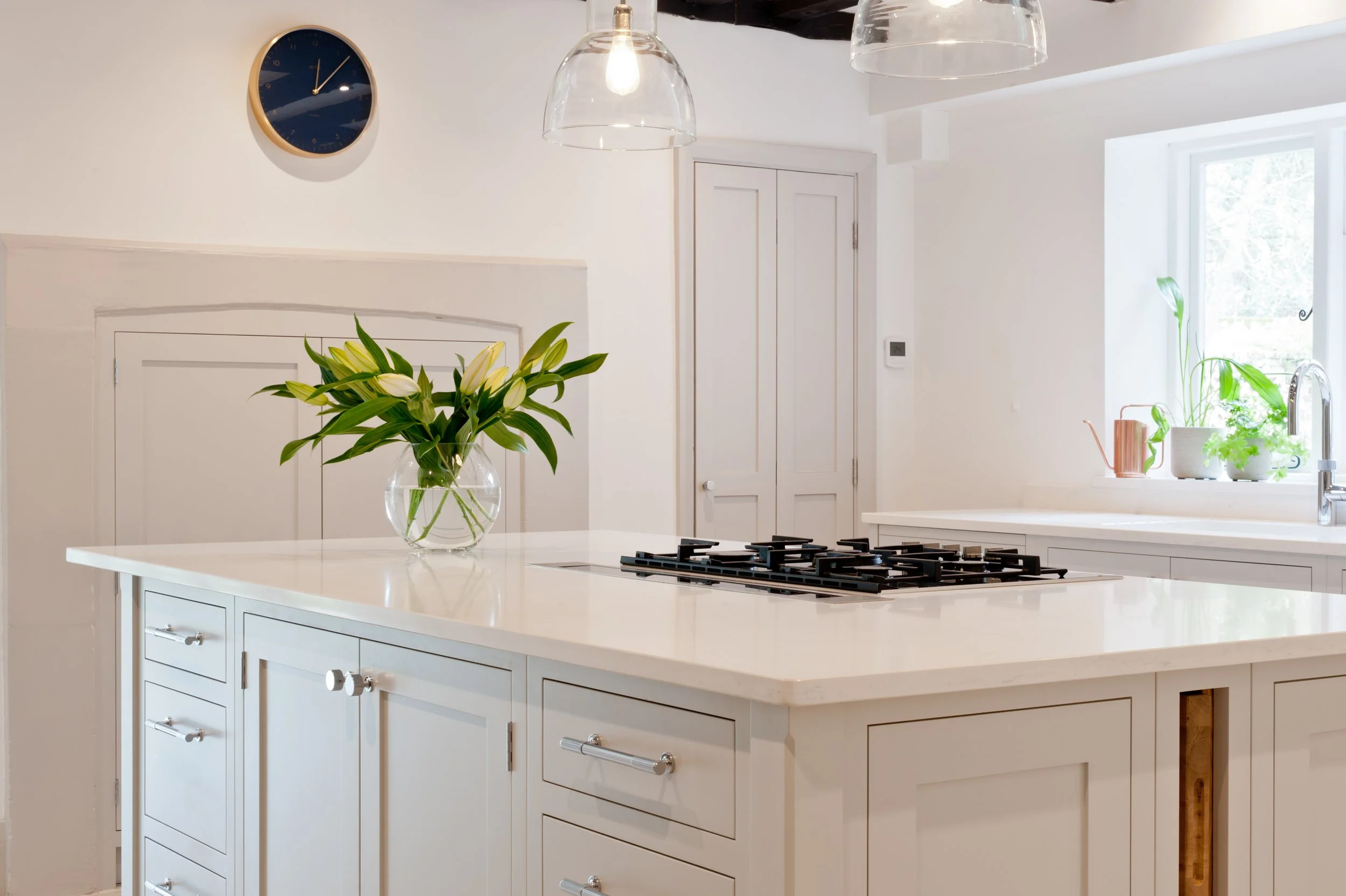 Bright kitchen with white cabinetry, a white island, a vase with white lilies on the island, and a large window with plants on the ledge. Modern pendant lights hang from the ceiling, and a wall clock is visible.