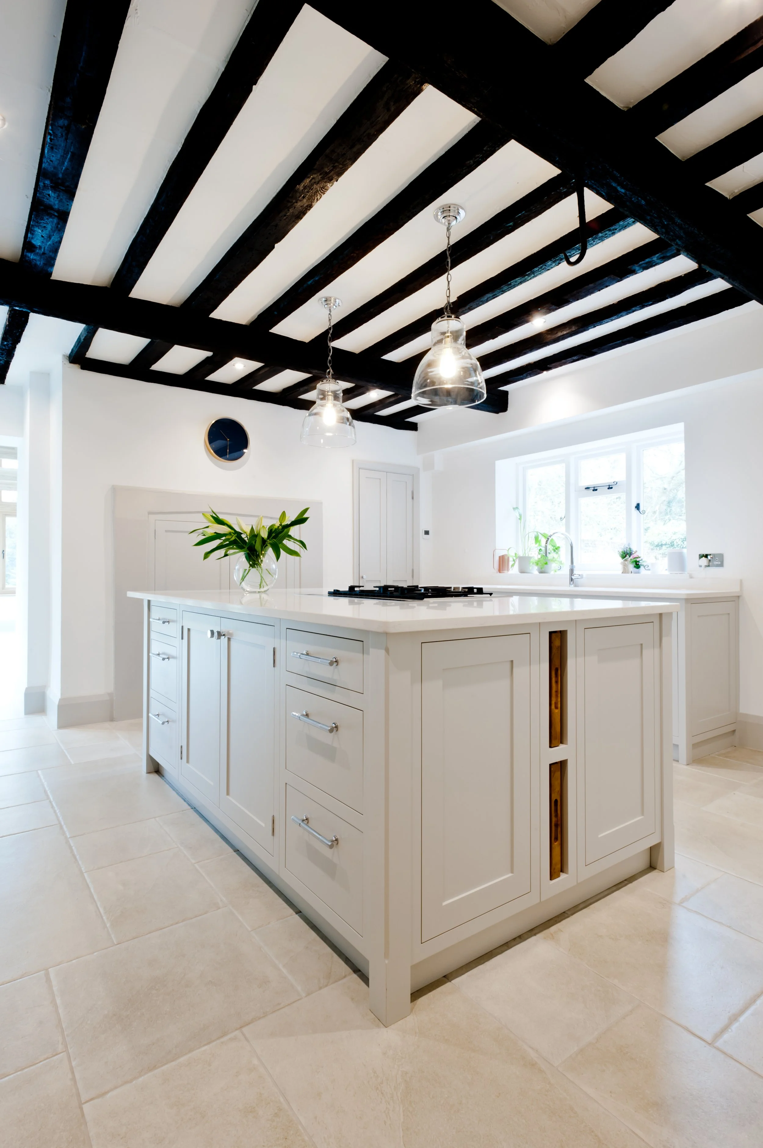 A white kitchen island with drawers, topped with a vase of white lilies, in a bright kitchen with black wooden ceiling beams, pendant lights, a window with plants, and a round wall clock.