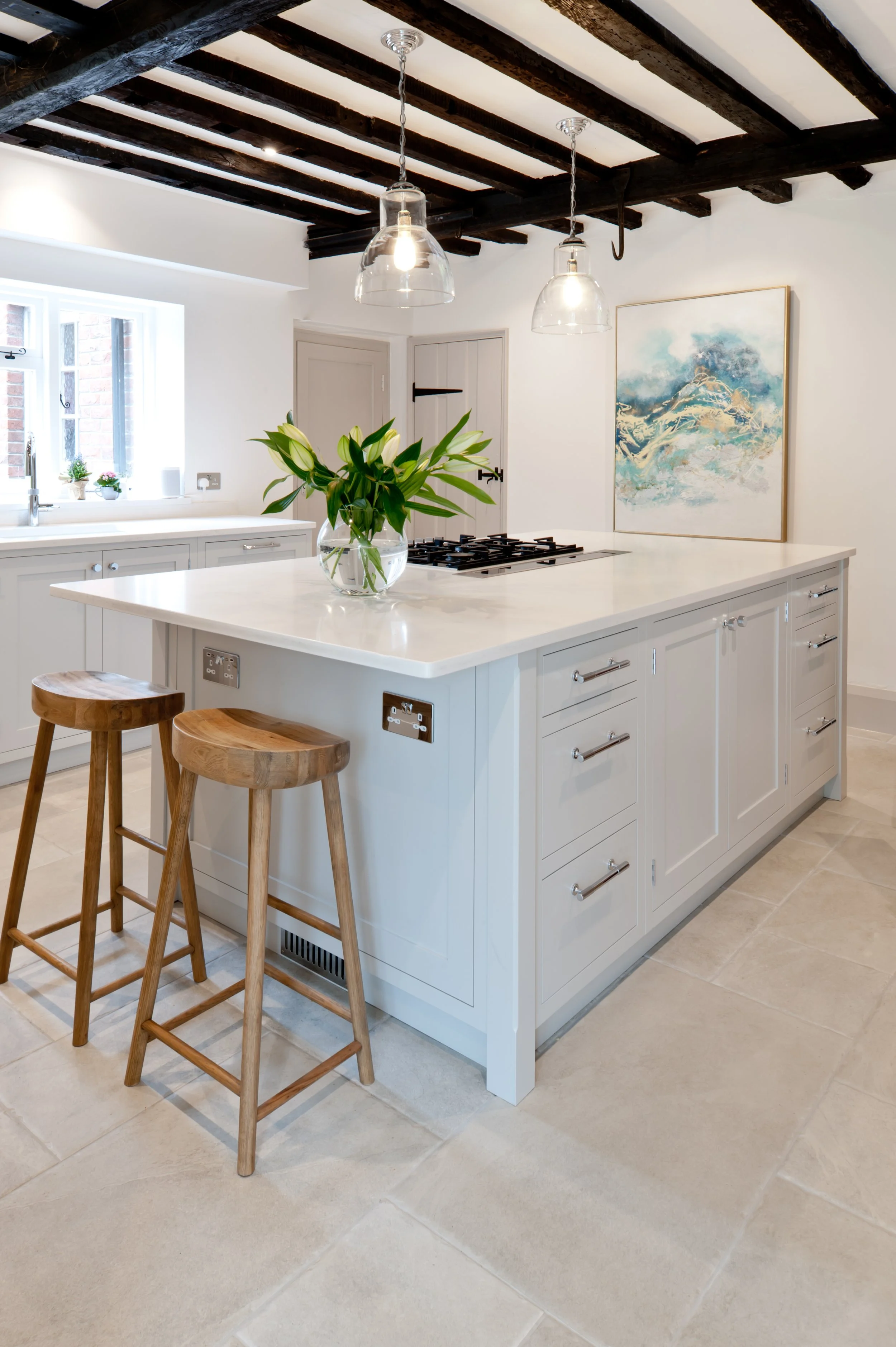A modern kitchen with white cabinets, a large white island, two wooden bar stools, a vase with white flowers on the island, a window with small potted plants, and a wall art piece with blue and beige tones. The ceiling has dark wooden beams and hangi