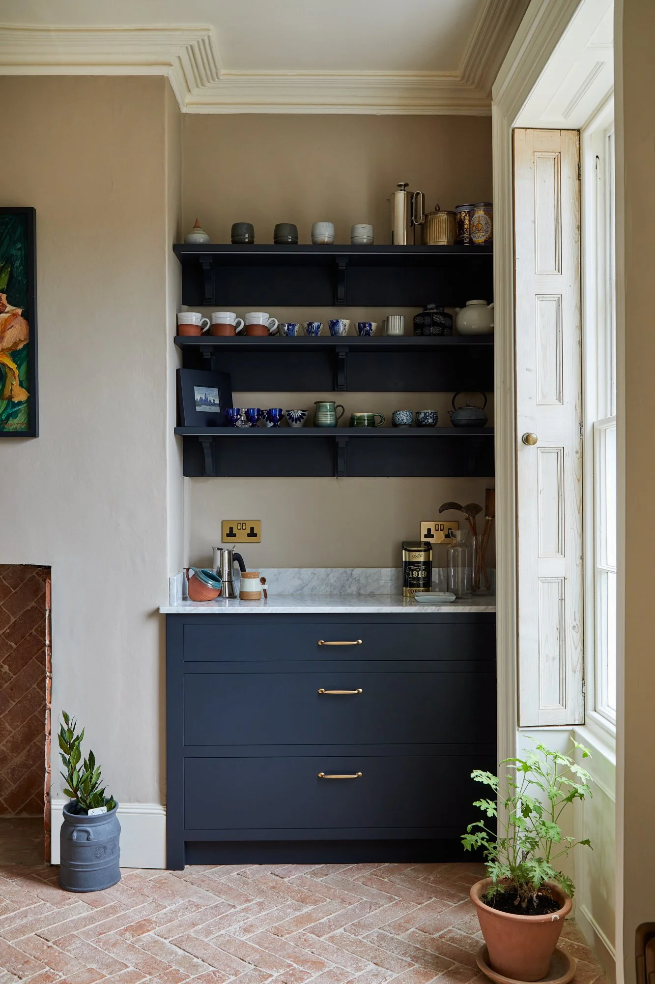 A kitchen corner with a navy blue cabinet and open shelving filled with cups, mugs, and jars. A marble countertop holds a coffee pot and cups. Two potted plants, one in a gray pot and the other in a terracotta pot, are on the brick floor. A window with open shudders allows natural light.