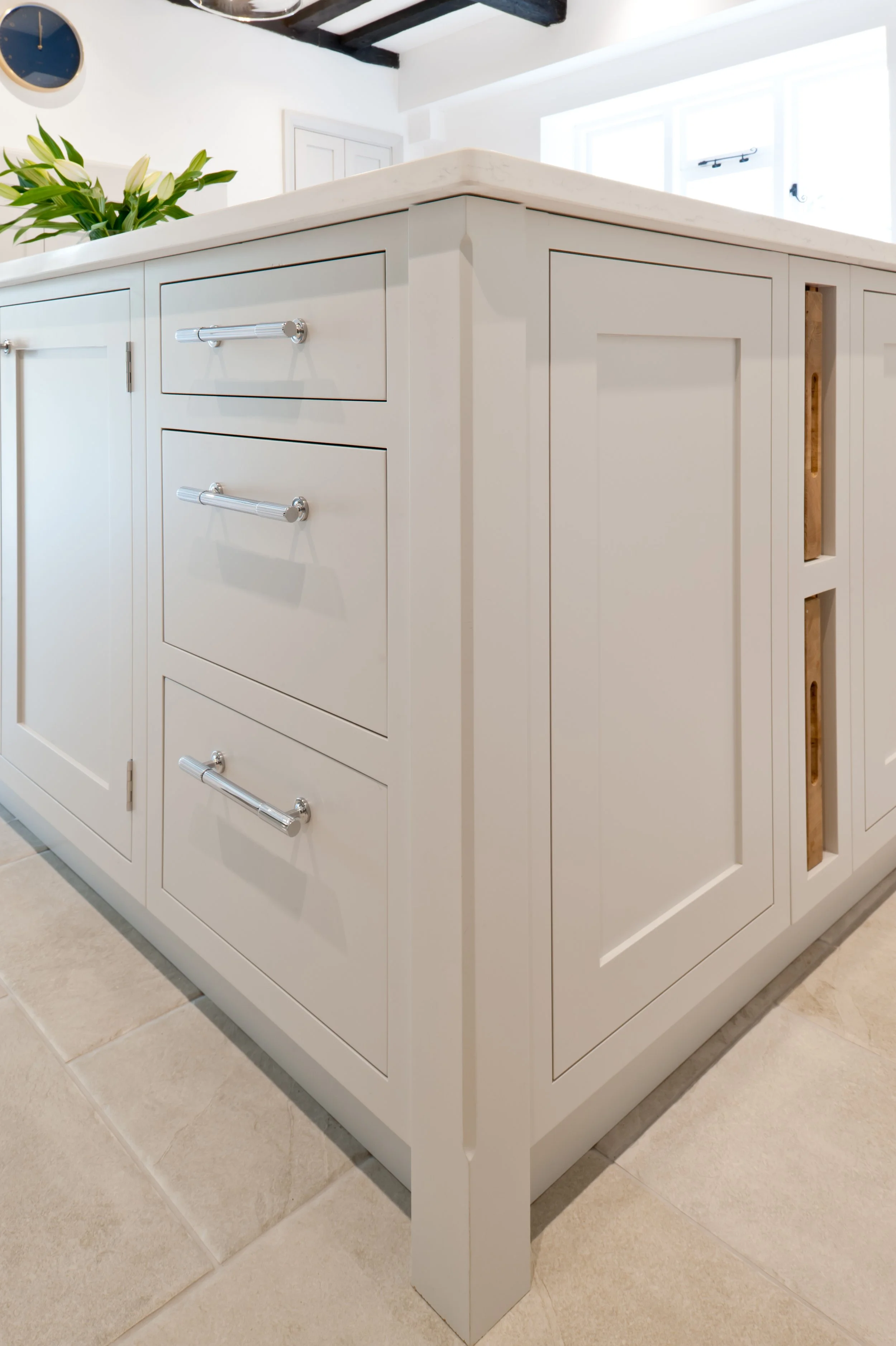 White kitchen island with six drawers and cabinets. The top is a smooth, light-colored stone. A potted plant with white flowers is on the island. The floor is tiled, and part of a window is visible in the background.