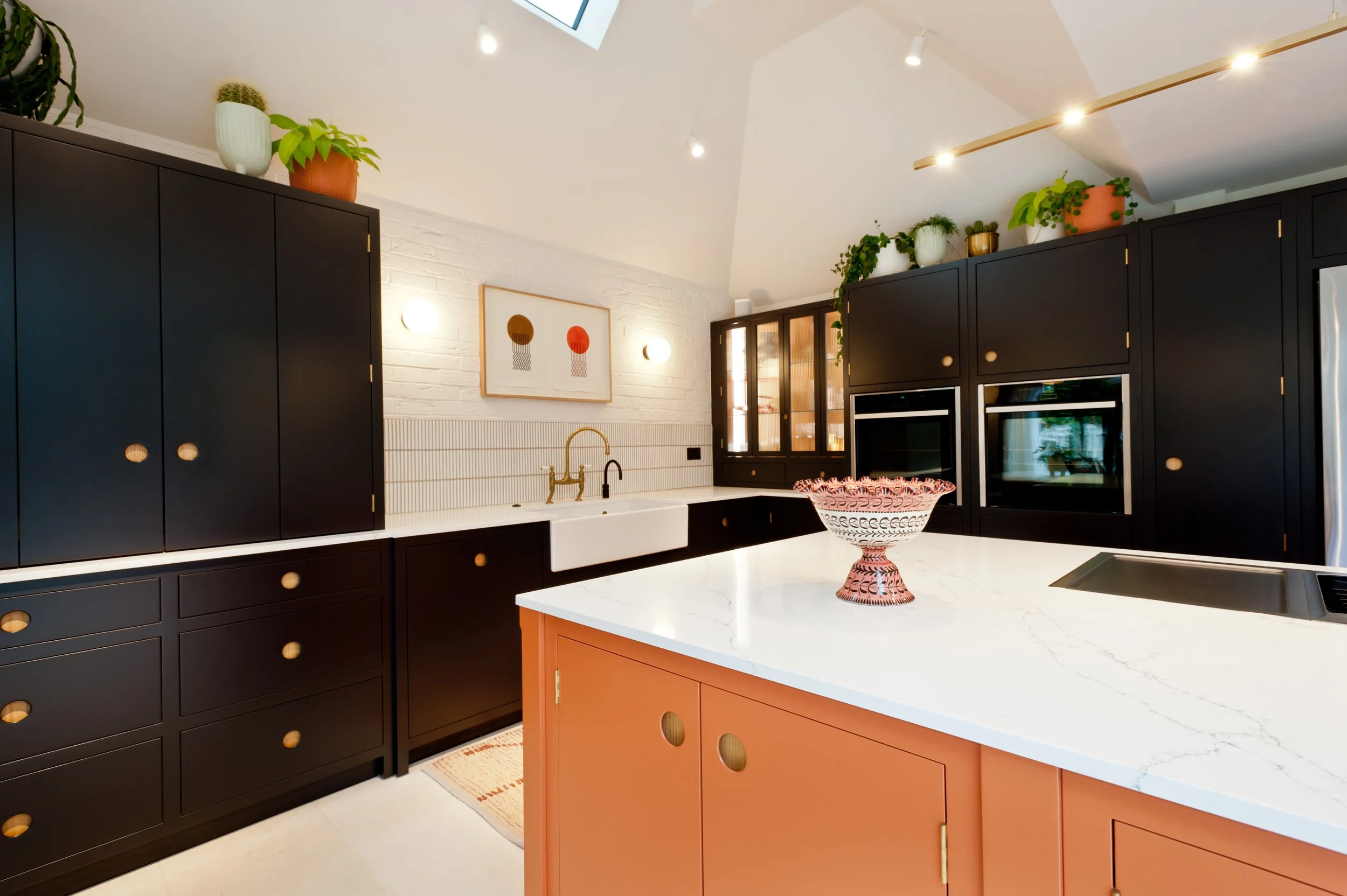 A modern kitchen with black and pink cabinets, white marble countertops, and decorative plants on top of the cabinets. There is a framed artwork, a white sink with a gold faucet, and a pink decorative bowl on the island, set under a sloped ceiling with a skylight and track lighting.