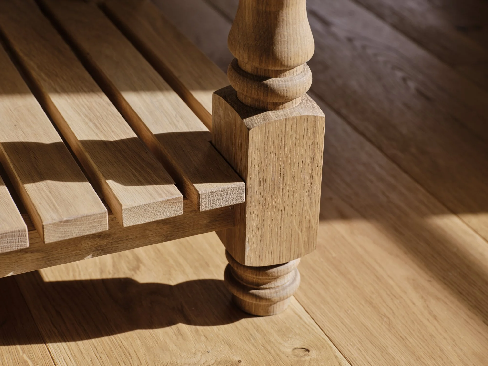 Close-up of a wooden bench with a detailed wooden post and slatted seat, on a wooden floor with sunlight casting shadows.