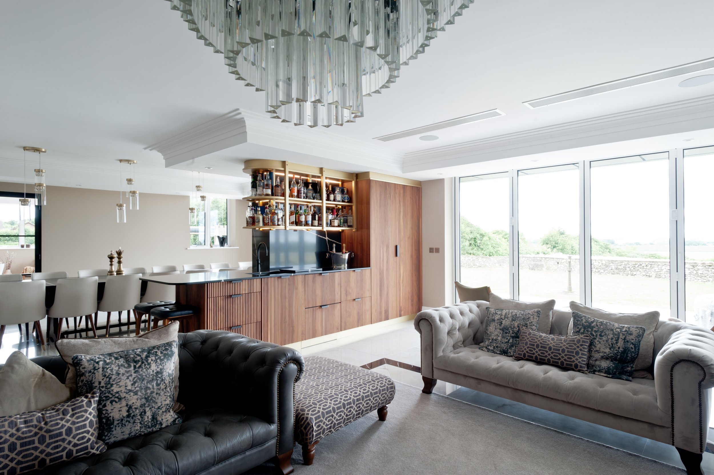 Living room with large sofa, accent pillows, and a chandelier, adjoining a kitchen with bar stools and a bar area with liquor bottles.