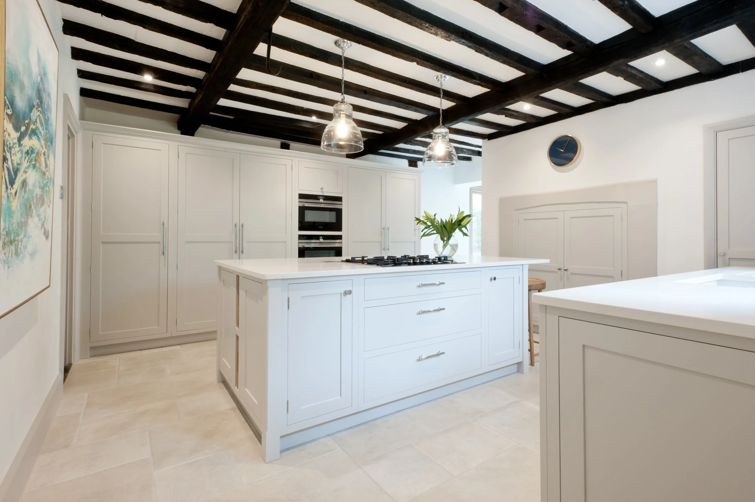 White kitchen with a large island, dark wood ceiling beams, modern pendant lights, white cabinetry, and beige tile flooring, with a vase of flowers on the island.