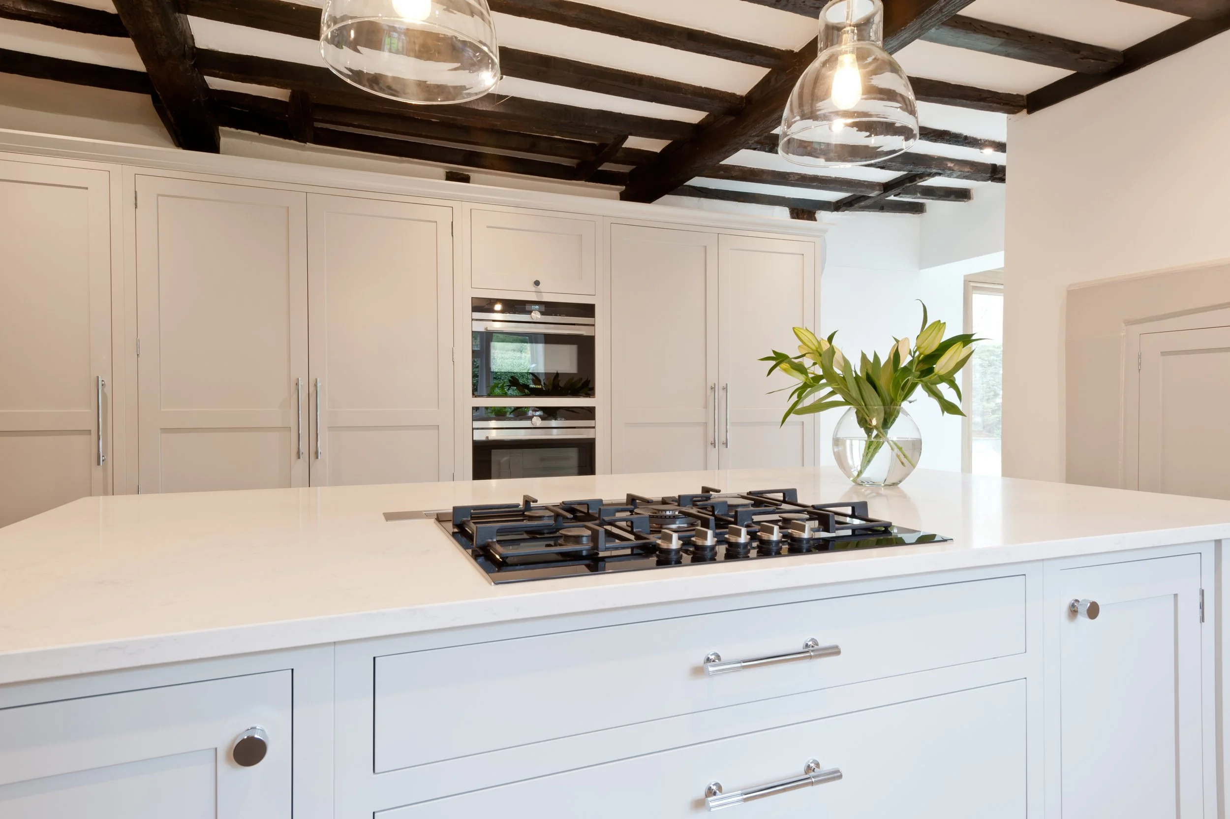 A modern kitchen with white cabinets, a white island counter with a built-in gas stove, a vase with white lilies, and pendant lights hanging from a dark wooden beamed ceiling.