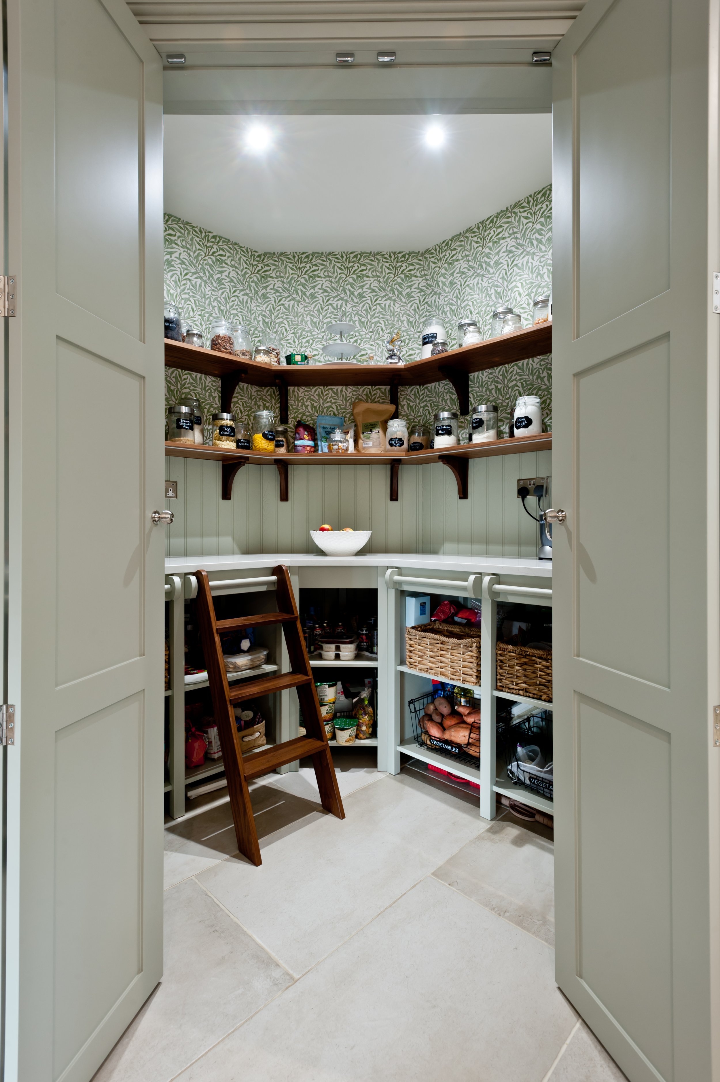 A pantry with double doors open showing shelves and countertop with various jars, baskets, and items like sweet potatoes and apples, with a small step stool inside.