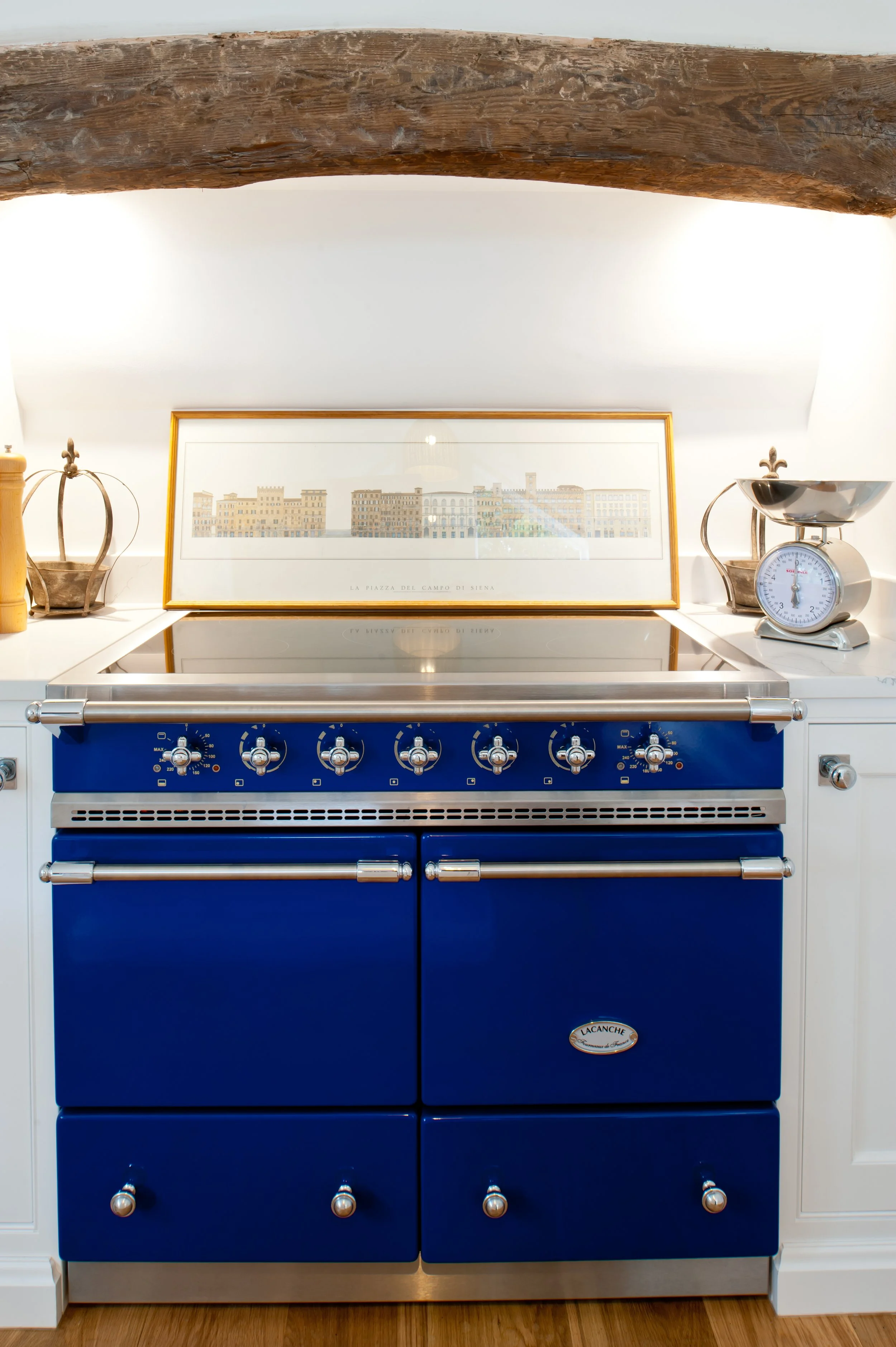 A vintage blue Lacanche kitchen stove with four burners and four oven drawers, positioned against a white wall with a framed cityscape print above it and decorative objects on either side.