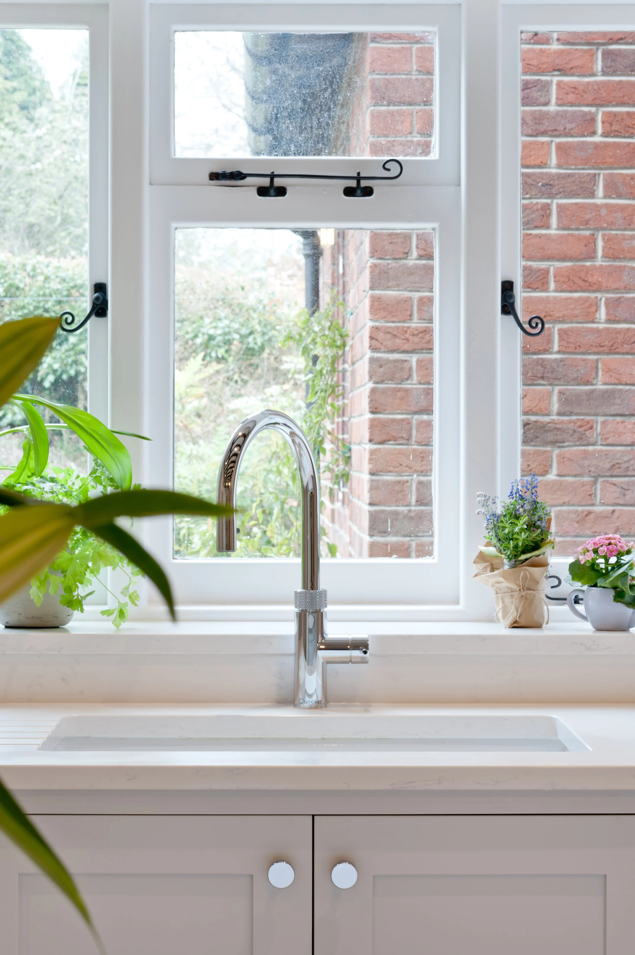 A kitchen window with a view of a brick wall and outdoor trees. The window has two sections with a central handle and decorative iron latch. Inside, on the windowsill, are potted plants, and a shiny kitchen faucet is in front of the window.