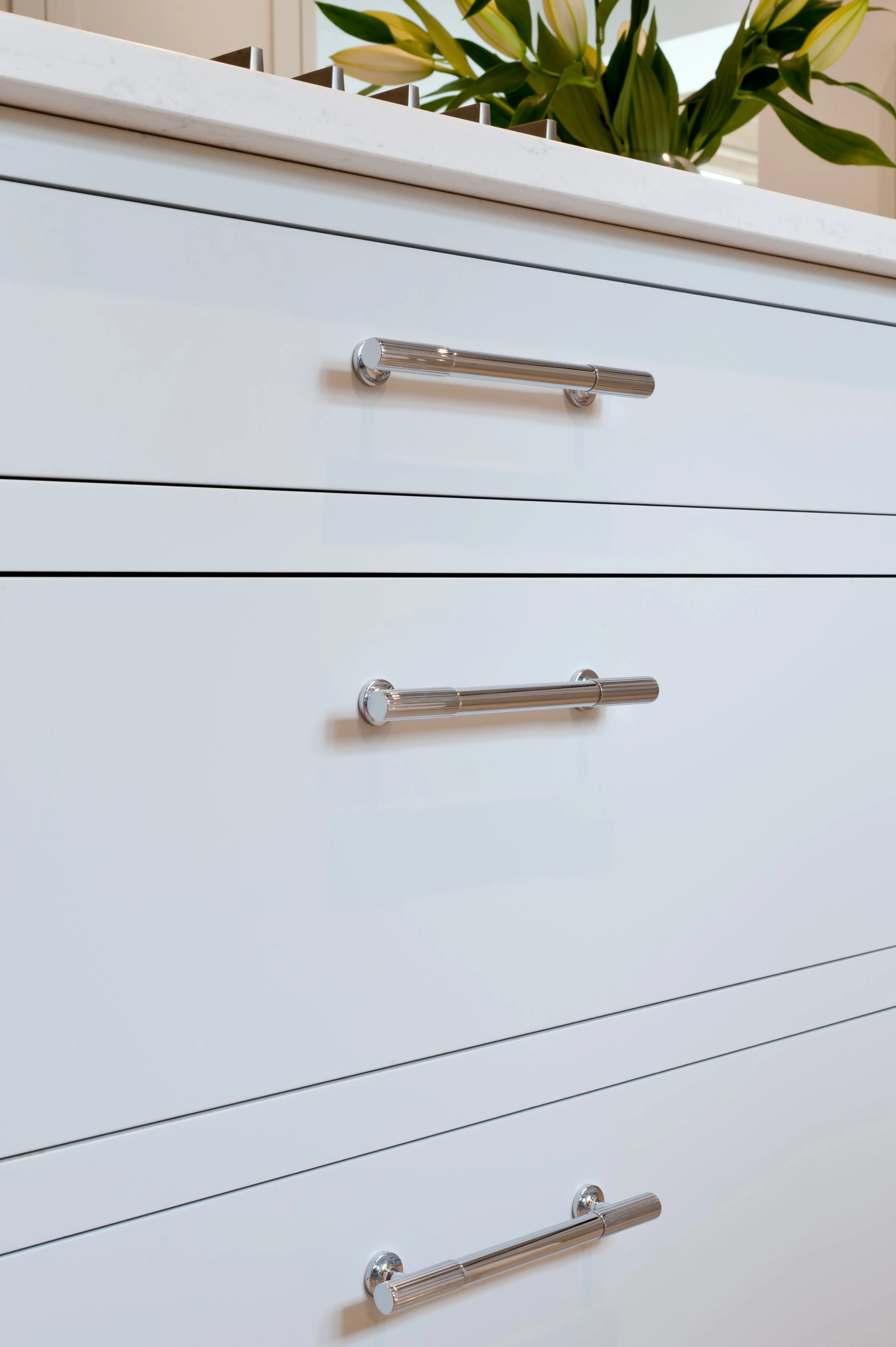 Close-up view of a white dresser with five drawers, featuring horizontal chrome handles. On top of the dresser, there is a rectangular planter with lush green leafy plants, placed on a white marble surface.