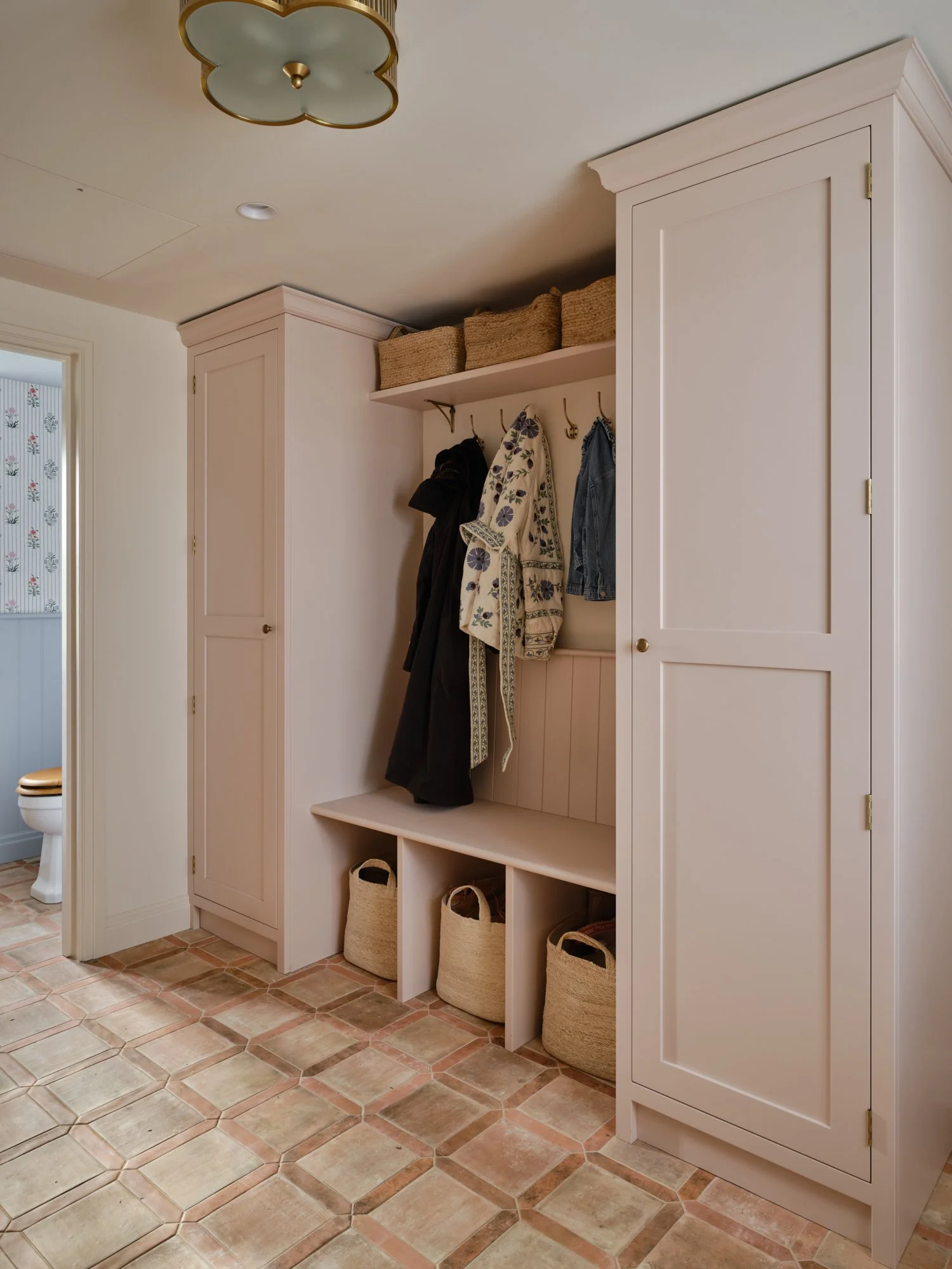 Bespoke boot room storage in pale pink with tall fitted cupboards, coat hooks, integrated bench seating and woven baskets beneath.