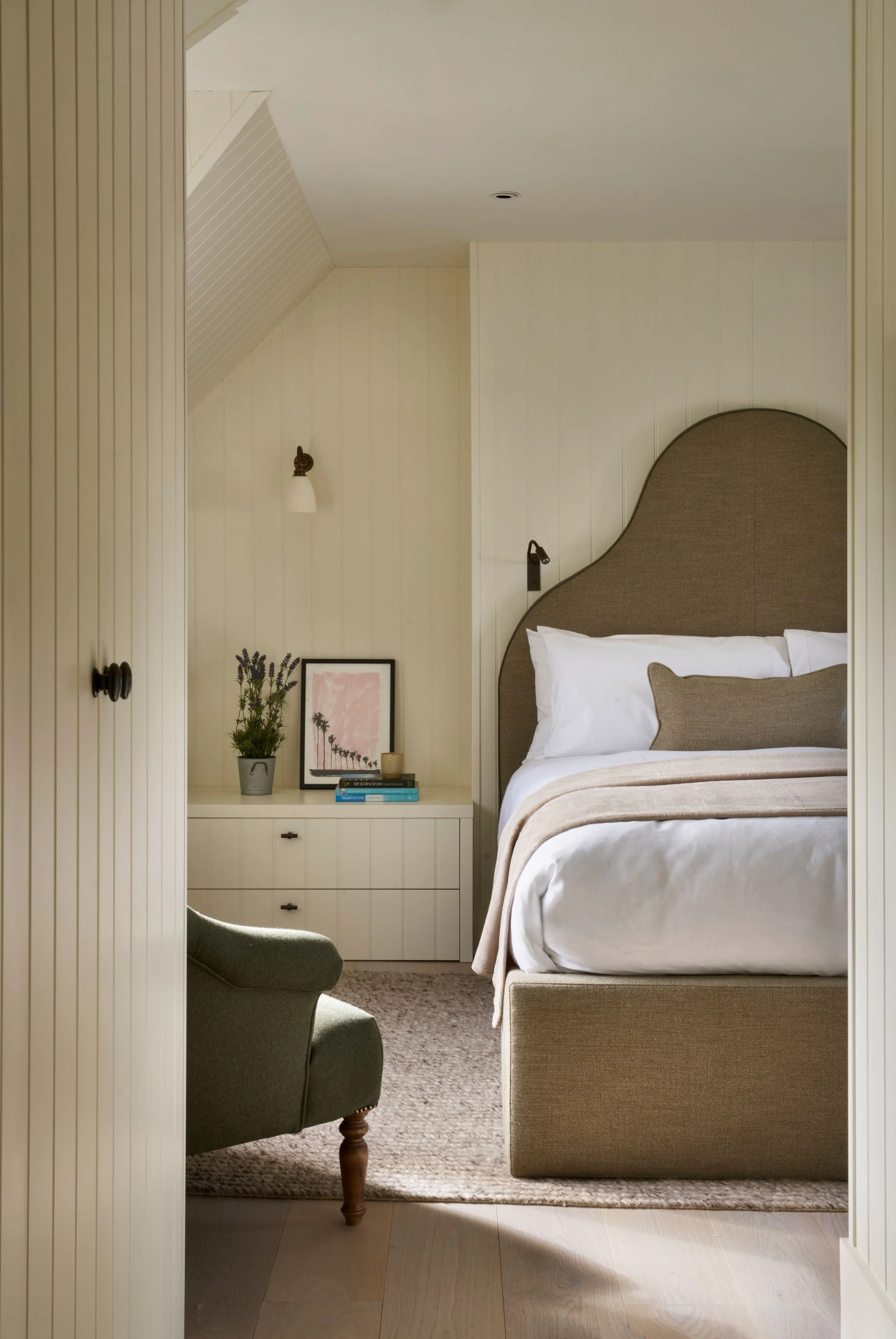 A cozy hotel bedroom with a bed, side table, artwork, and an armchair, seen through a doorway with wood paneling.