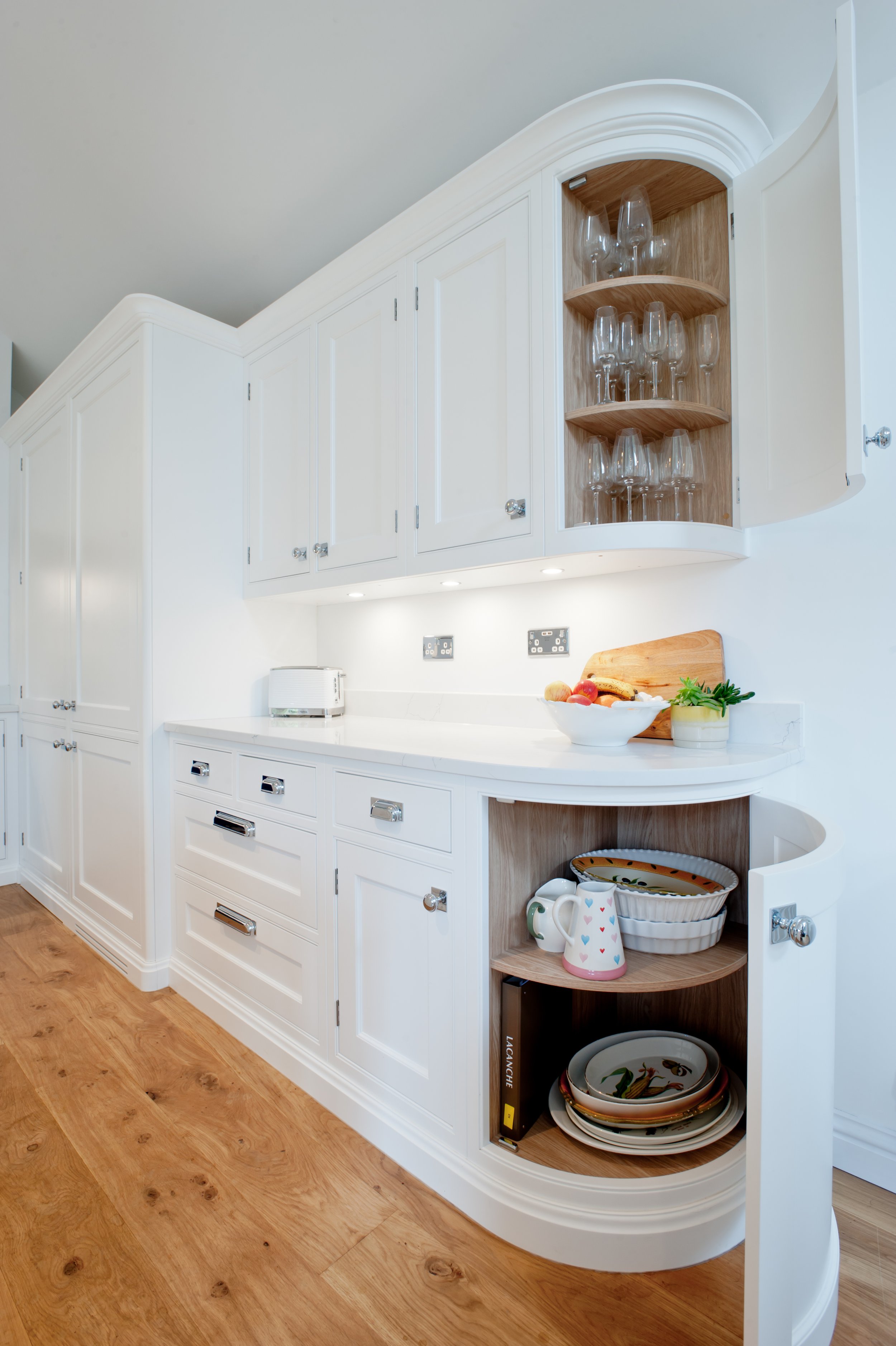 White kitchen cabinetry with open corner shelves holding glassware, a white countertop with a bowl of fruit, a cookbook, and decorative plates on a lower shelf, with a wooden floor.