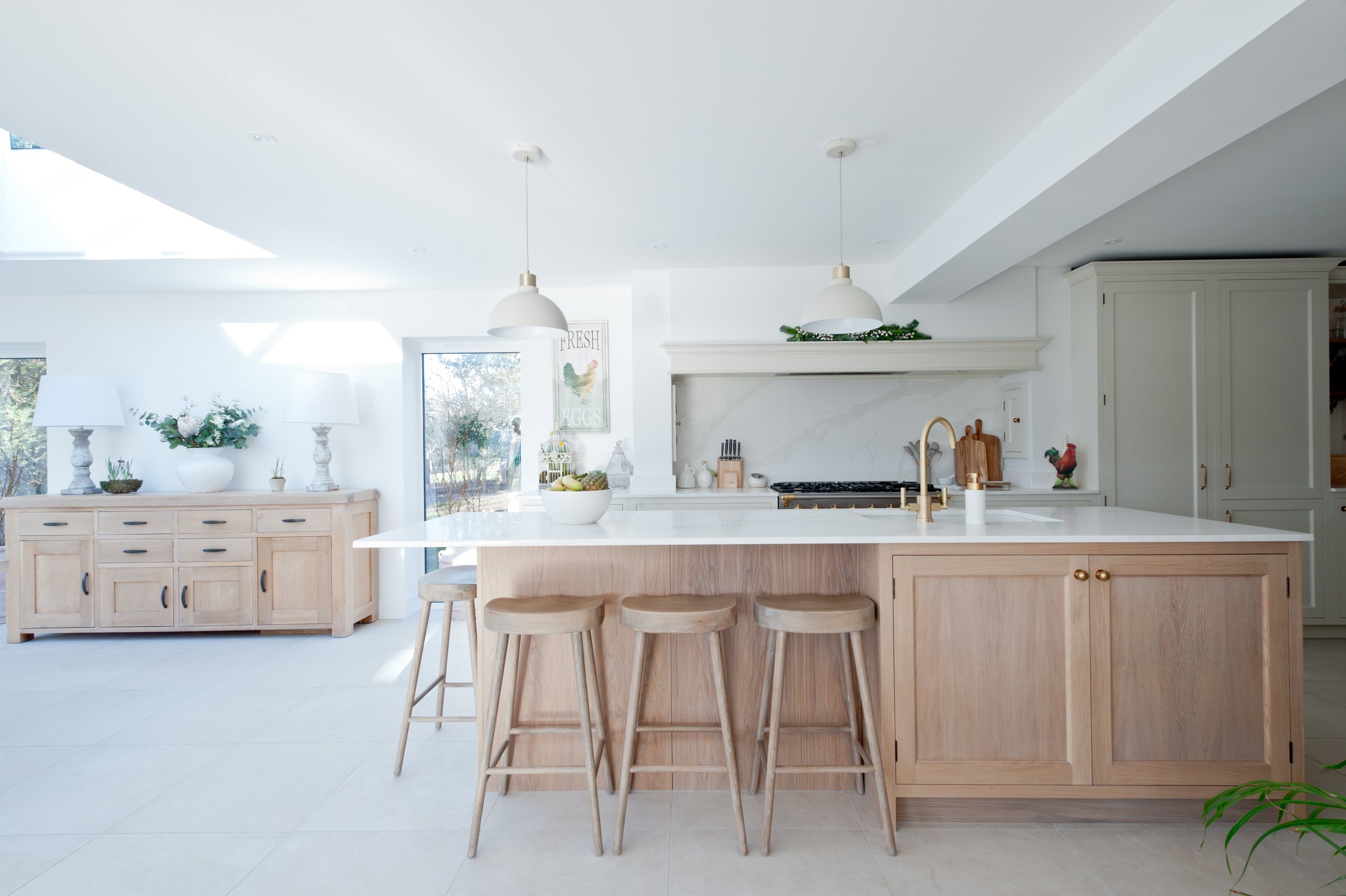 A modern kitchen with white walls and light wood cabinetry. The kitchen island has a white countertop with three wooden stools in front. There are two white pendant lights hanging above the island. Decor includes a bowl of fruit, a small potted plant, and a soap dispenser. A decorated garland sits on top of the range hood, and a rooster figurine is on the counter. Large windows on the left provide natural light.