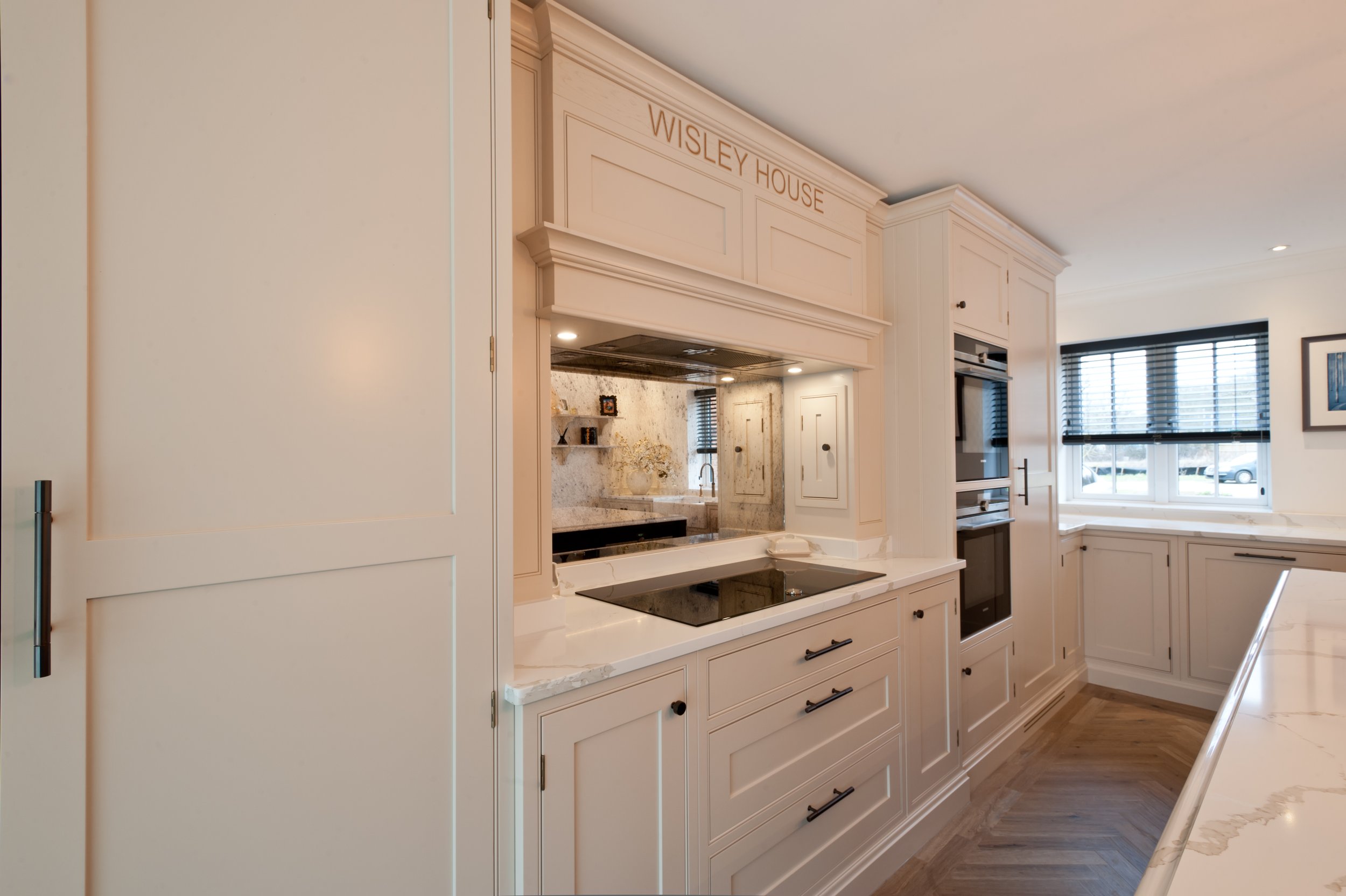 A modern kitchen with white cabinetry and black handles, a marble countertop with an electric cooktop, and a window with black blinds. The sign reads "WISLEY HOUSE."