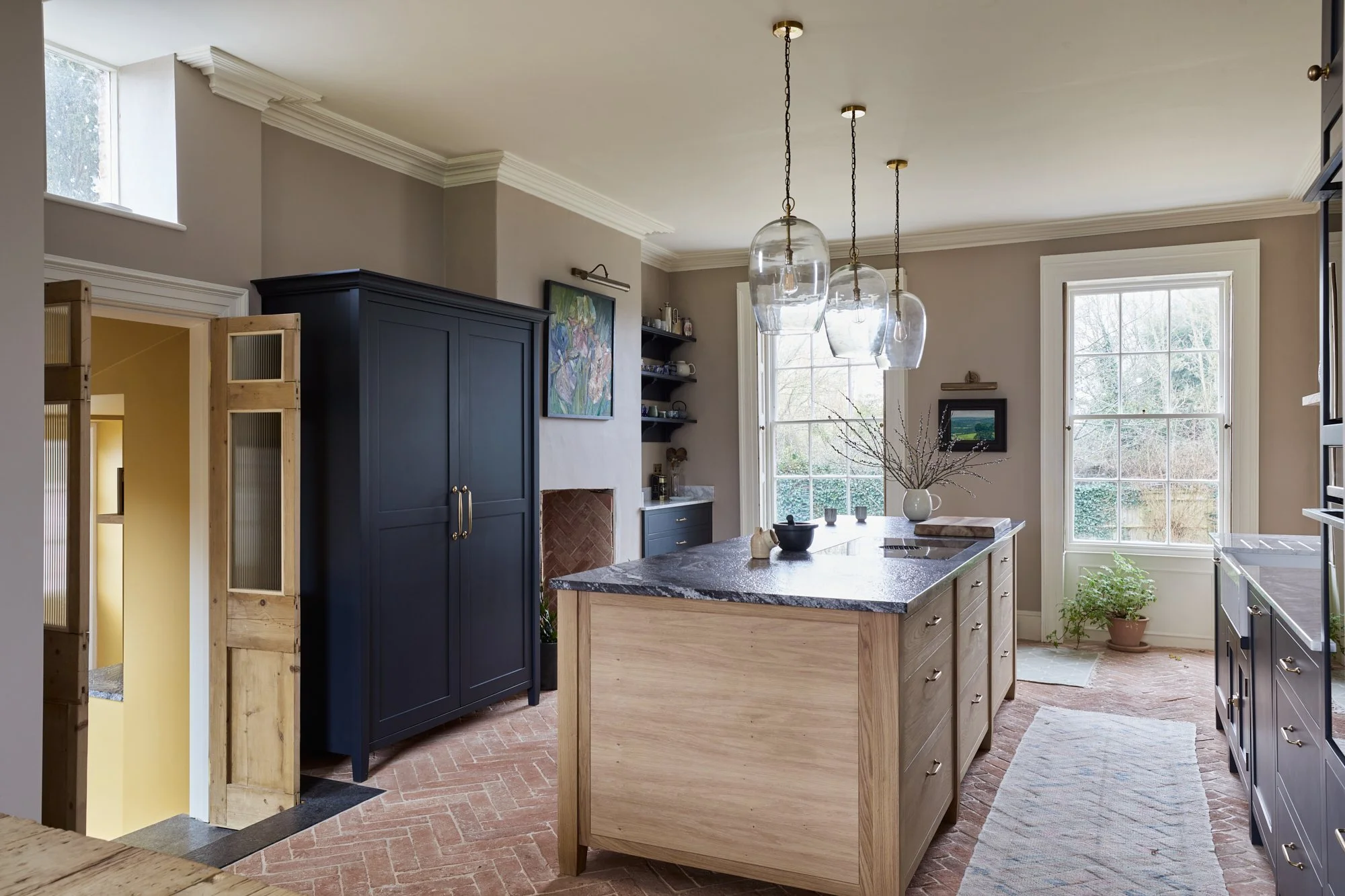 A cozy kitchen with large windows, a central island with a dark marble countertop, navy blue cabinetry, open shelving, and natural light.