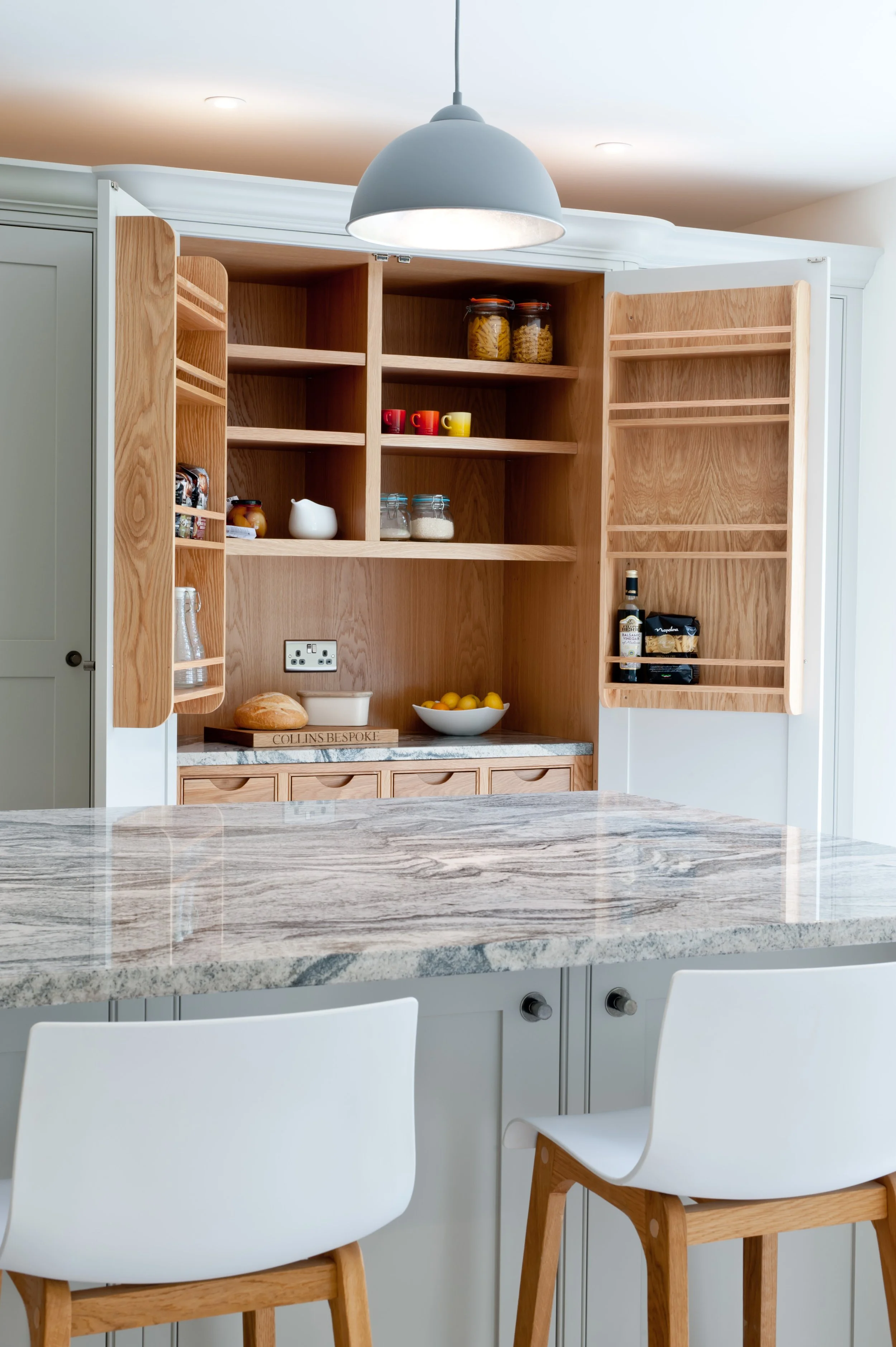 A modern kitchen with a marble countertop, white and wooden chairs, and open wooden cabinets with jars, mugs, and a few food items inside.
